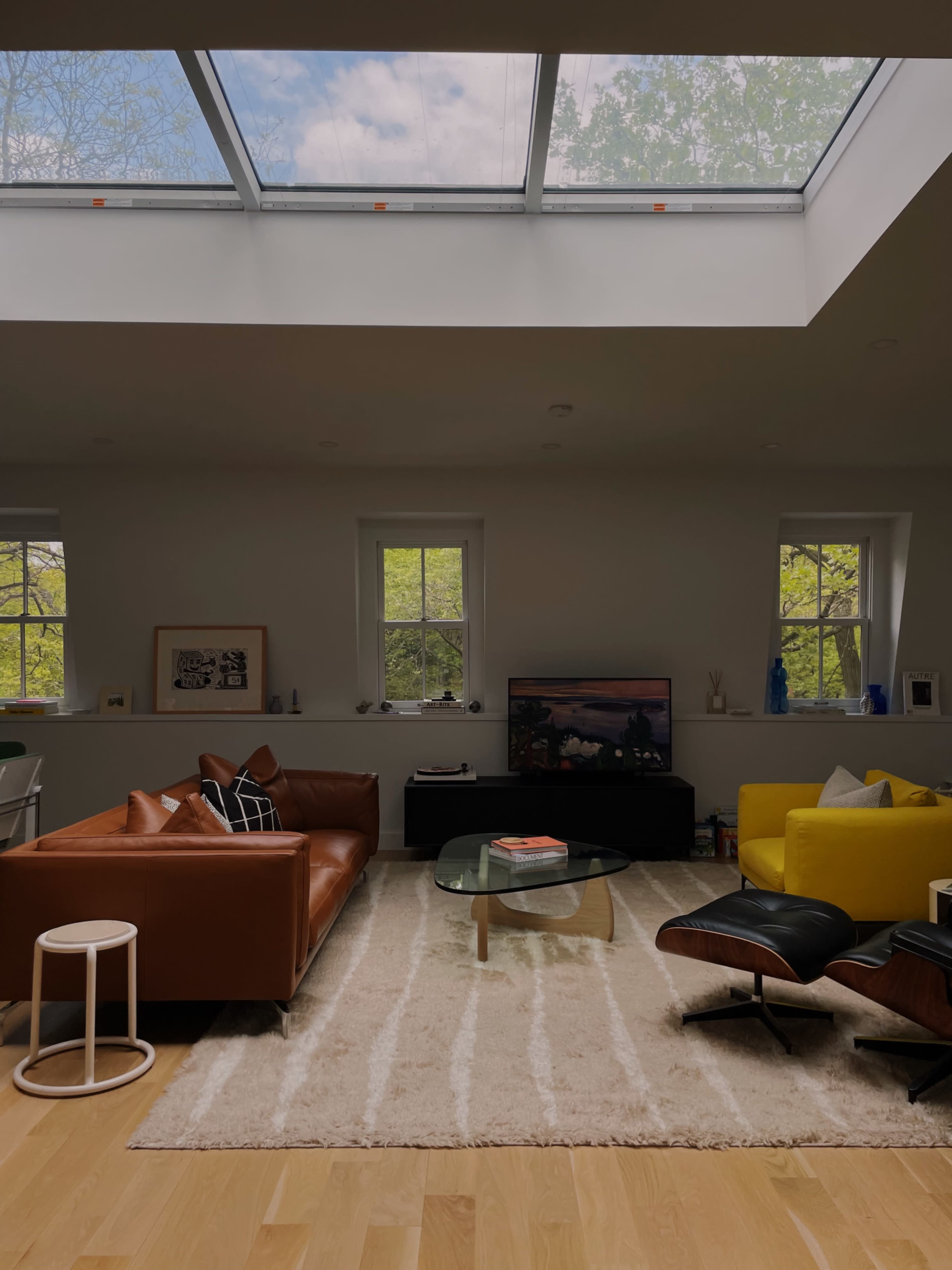 A cozy living room features a brown leather sofa, a yellow chair, and a glass coffee table, illuminated by natural light from a skylight.
