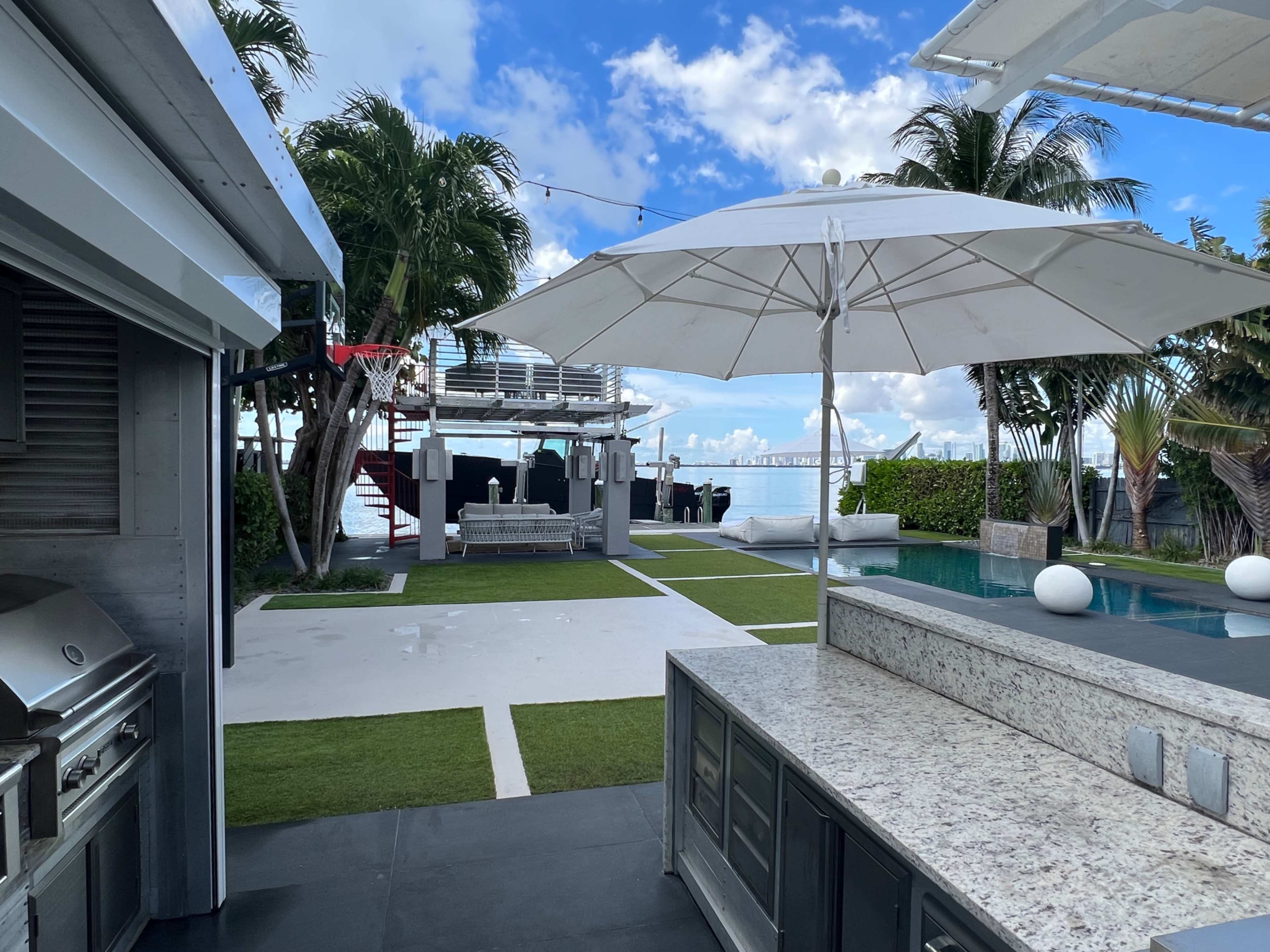 The image shows a modern outdoor kitchen with a granite countertop, an umbrella, and a view of a pool and skyline over the water, surrounded by palm trees and landscaped grass.