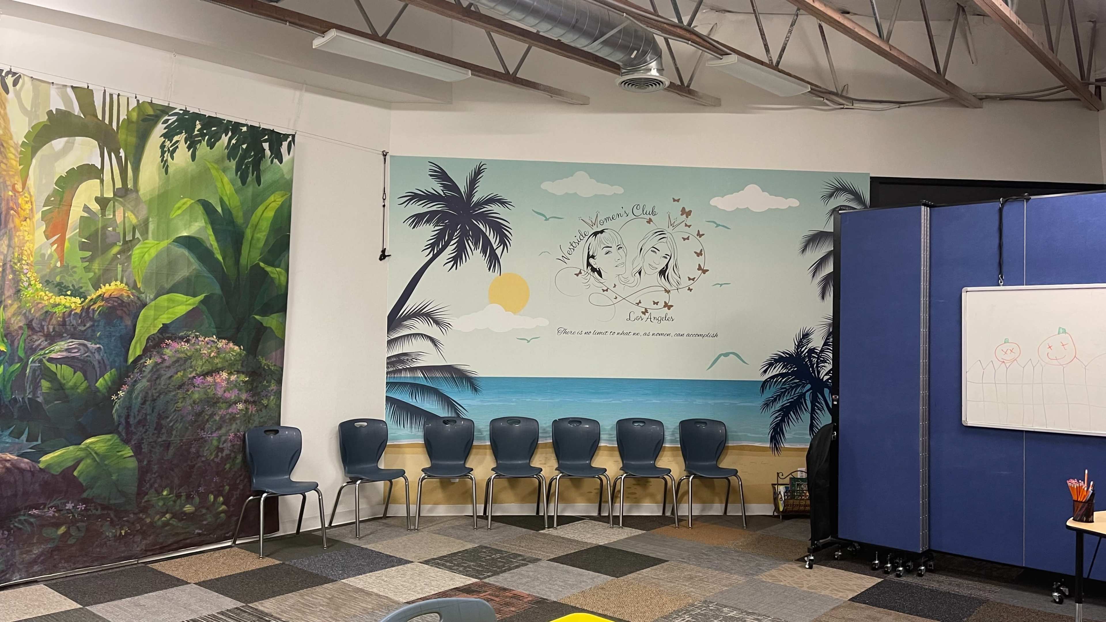A classroom with a mural of palm trees and ocean on one wall and paintings of greenery on another, with several blue chairs arranged in a row.