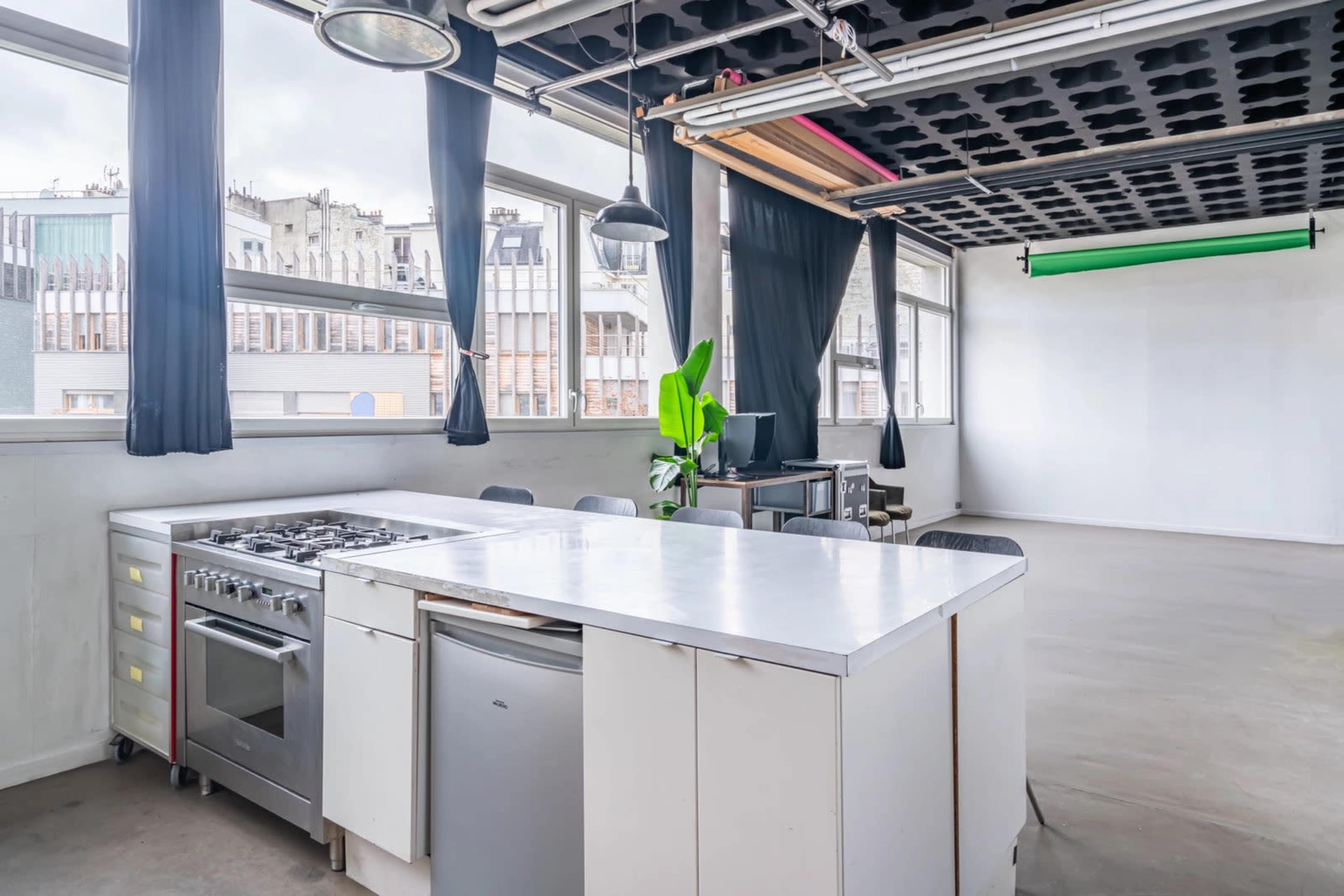 A spacious room features a central kitchen island with a gas stove and a refrigerator, surrounded by large windows with views of neighboring buildings.