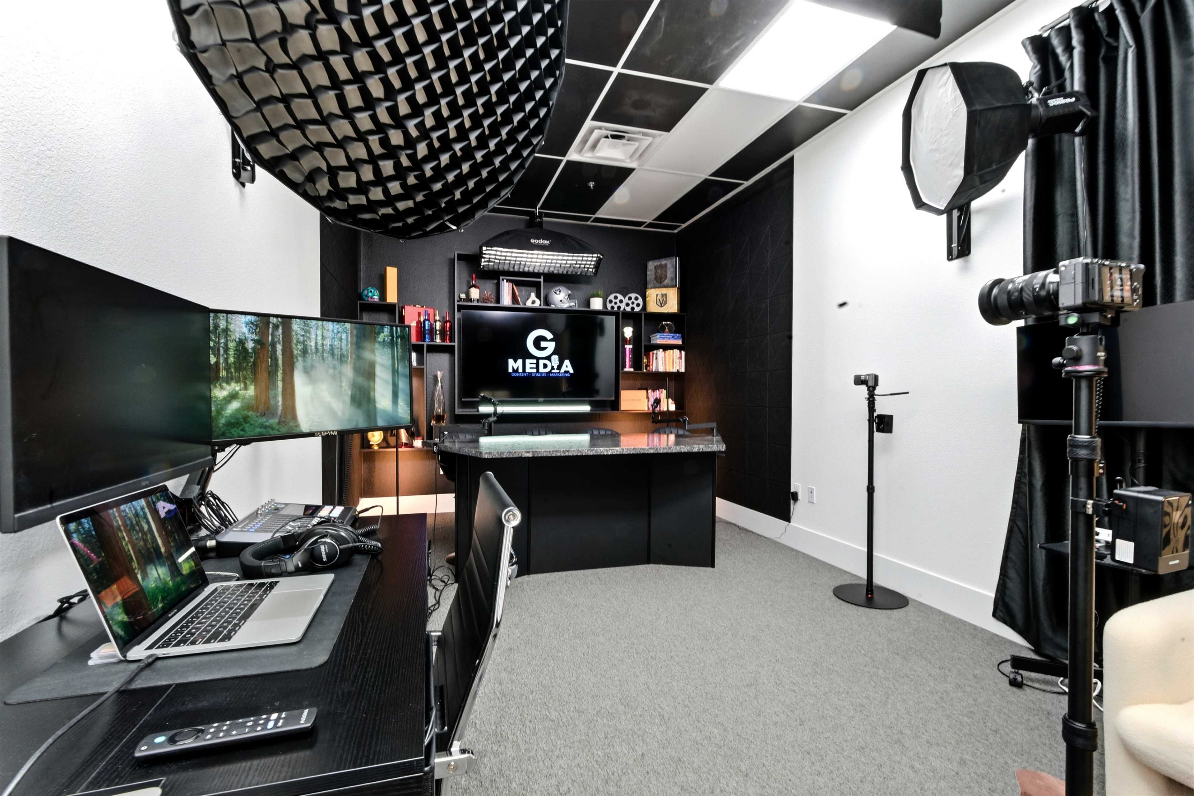 The image shows a modern media studio featuring dual computer monitors, a desk with a laptop, and professional lighting equipment arranged in a carpeted room.