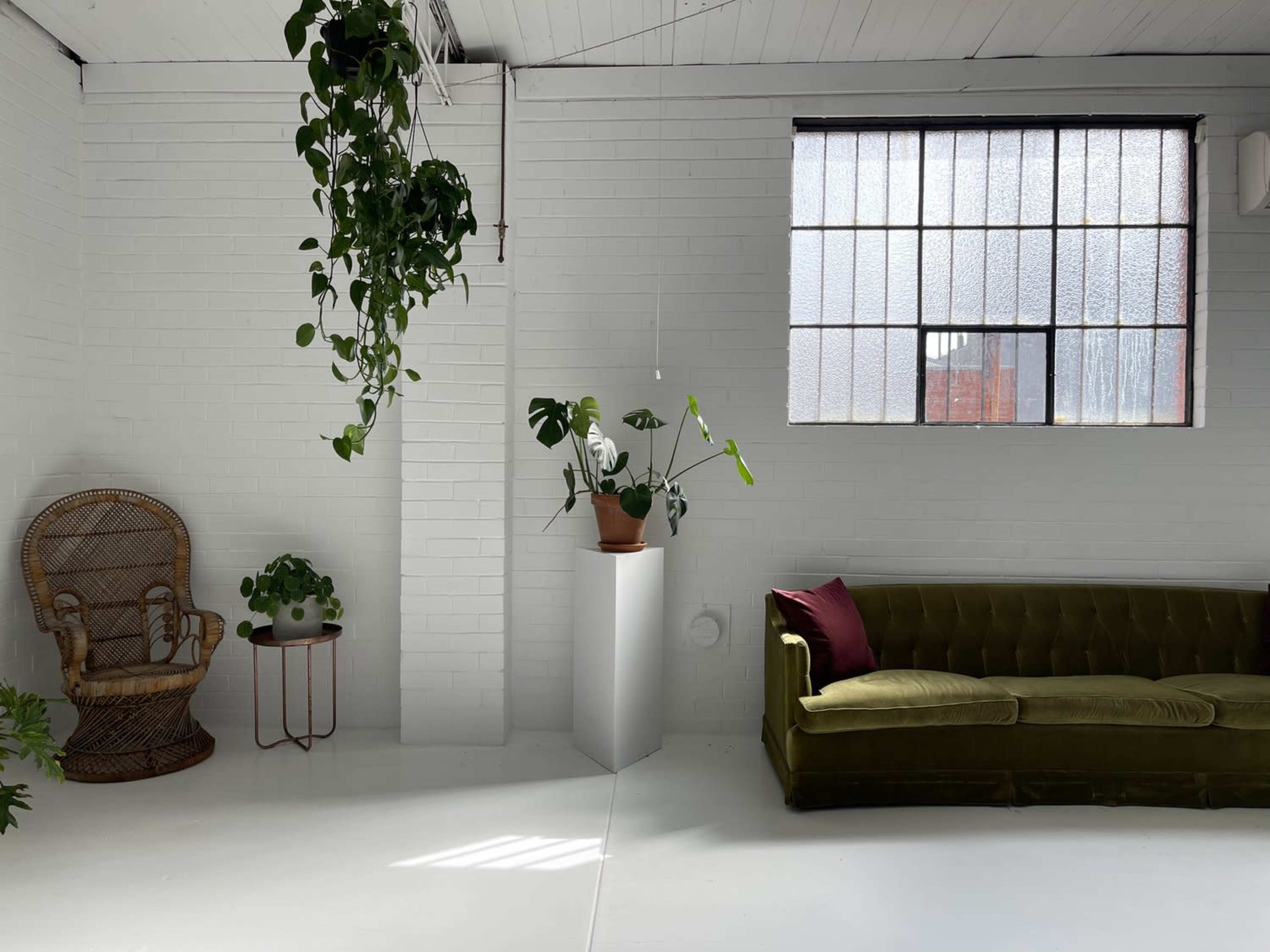 A green velvet sofa sits against a white brick wall beside a potted plant on a pedestal, with a rattan chair in the corner and large windows letting in natural light.