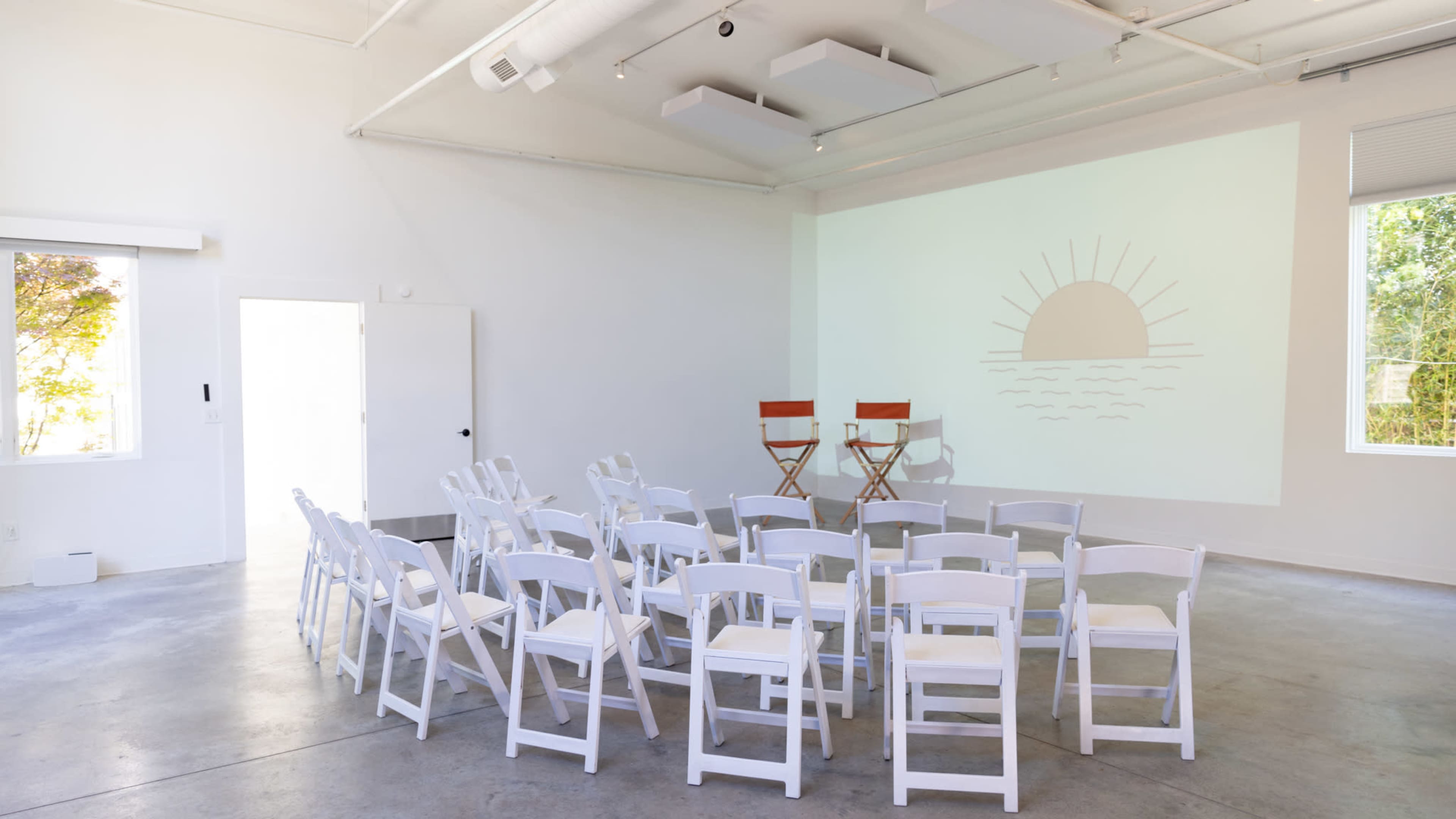 A spacious room with white walls and a simple projection of a sun design on one wall, arranged with circle seating of white chairs and two director's chairs at the front.