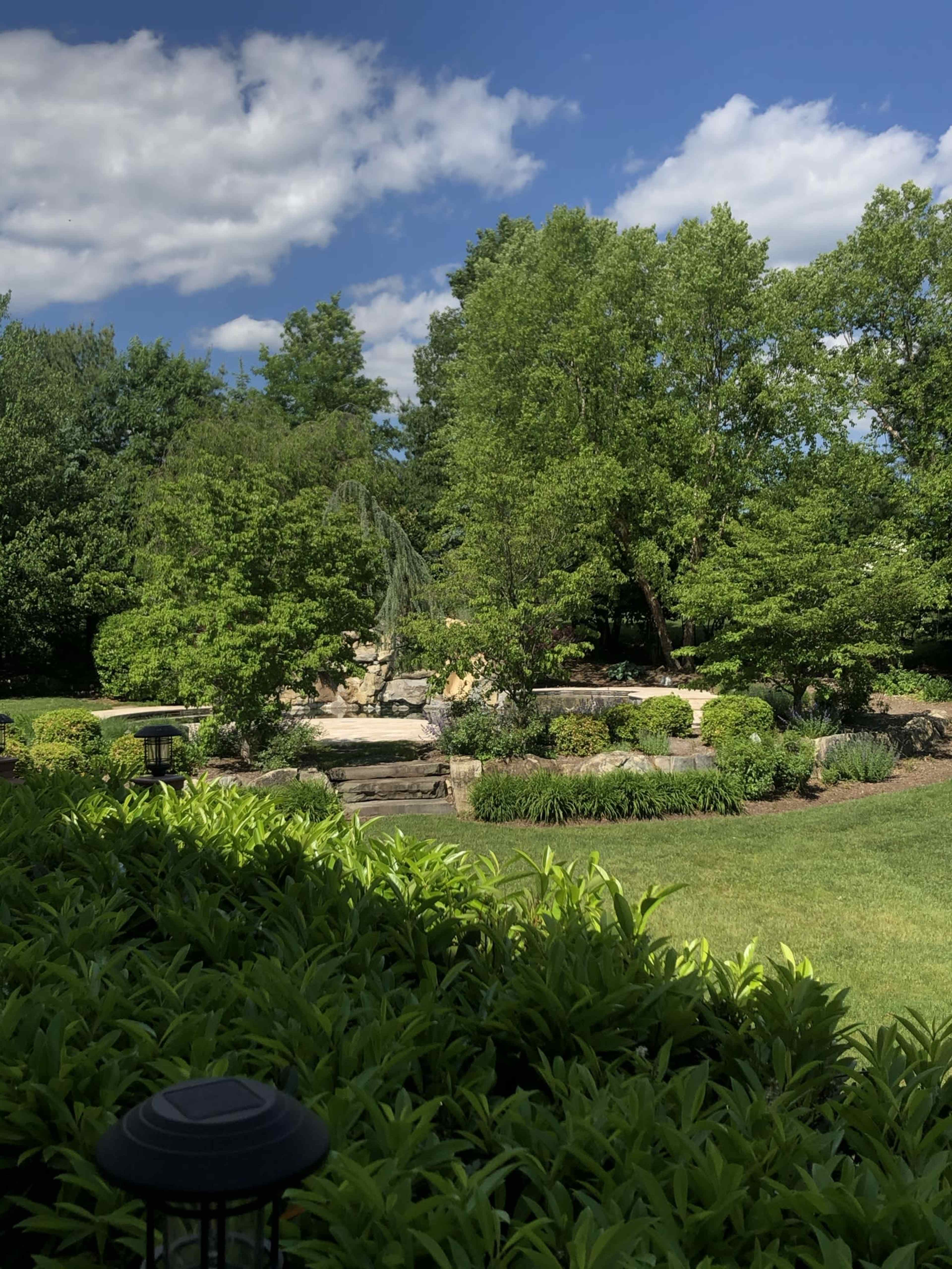 A landscaped garden featuring a rock fountain surrounded by lush greenery and trees under a partly cloudy sky.