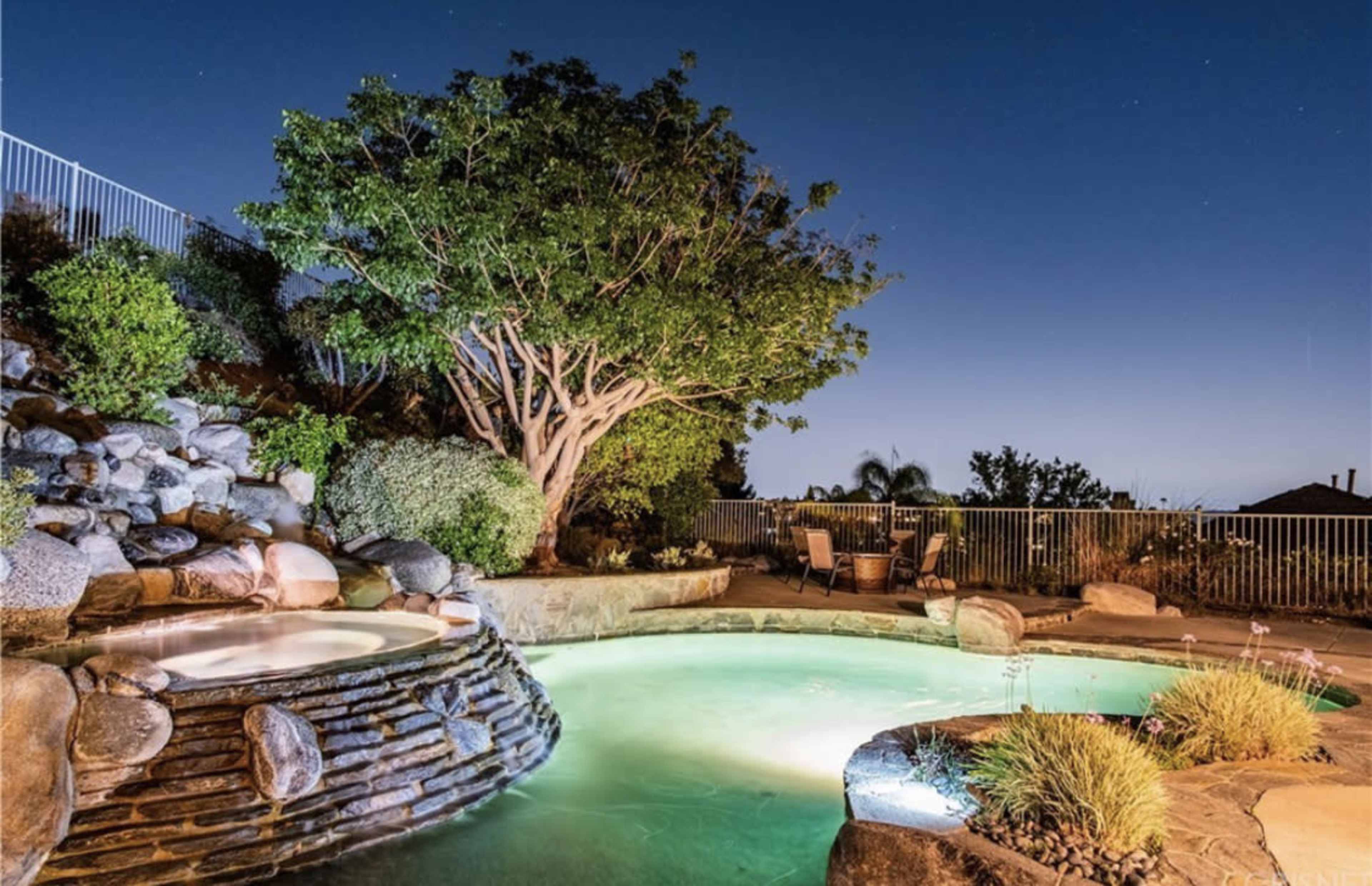 A illuminated backyard features a stone hot tub beside a pool and a large tree at dusk.