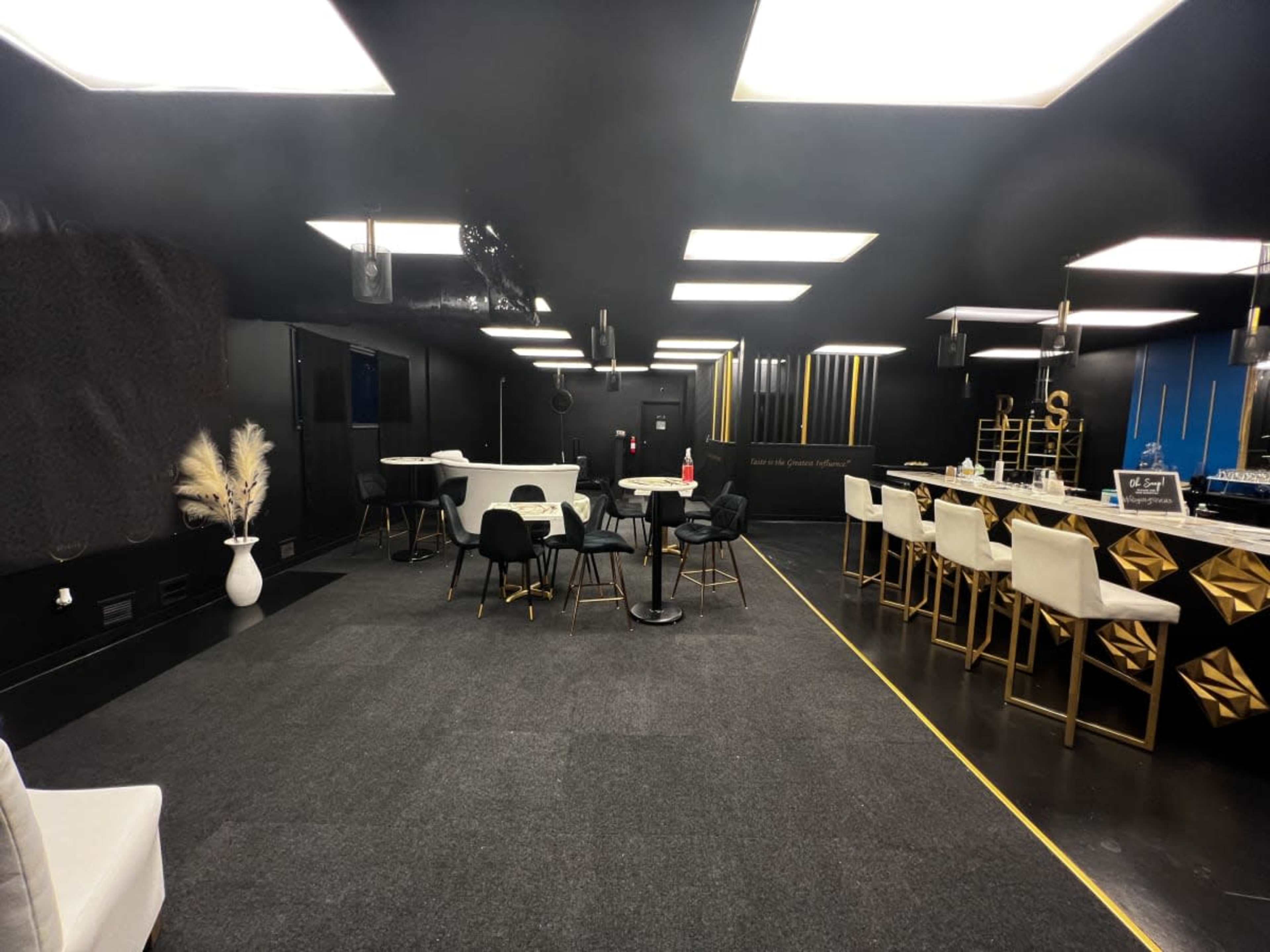 A modern, dimly lit lounge area with black walls, gold accents, tables, and chairs arranged around a bar with high stools.