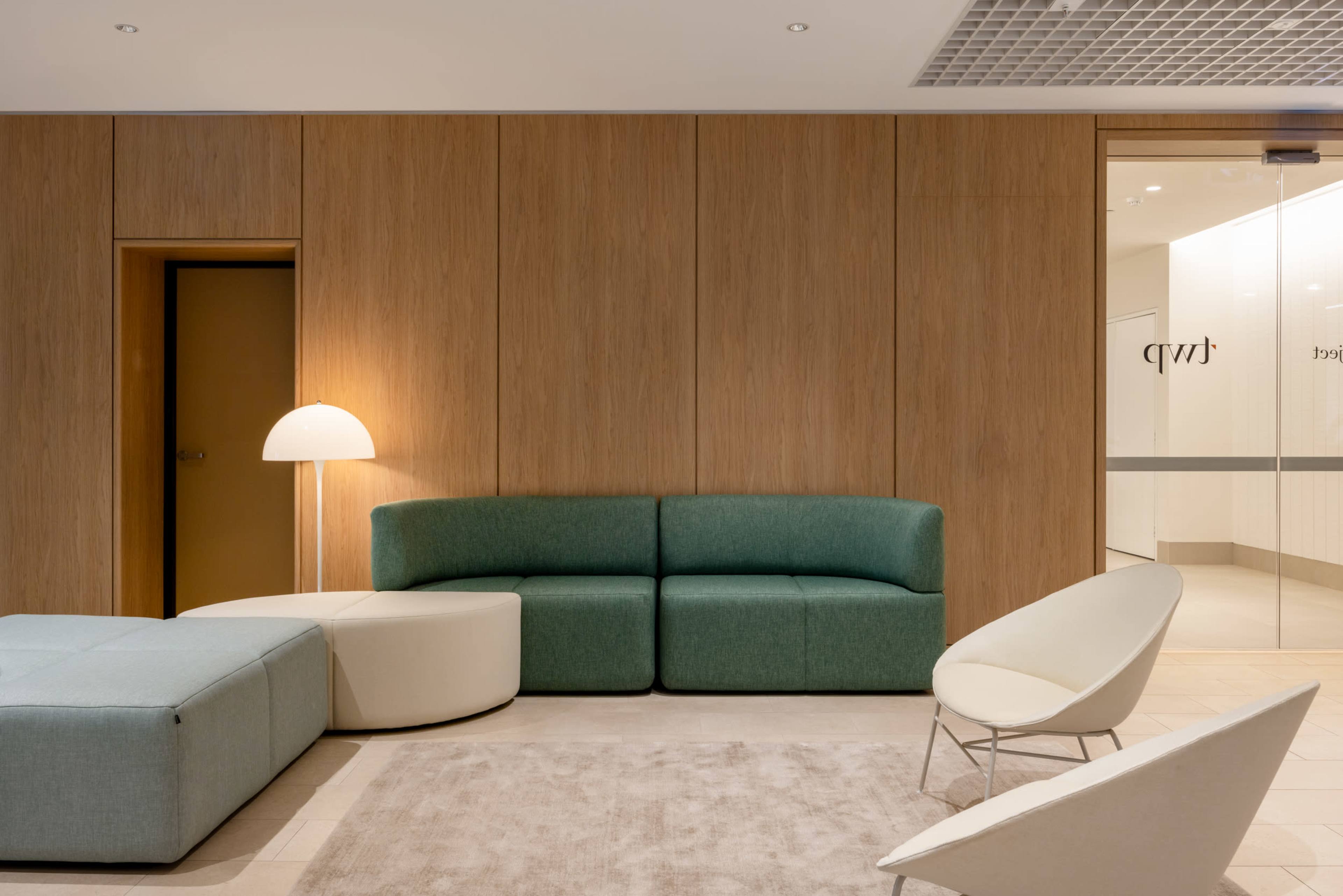 The image shows a modern waiting area featuring a green upholstered sofa, a circular white table, a floor lamp, and a light-colored accent chair, all set against a wooden-paneled wall.