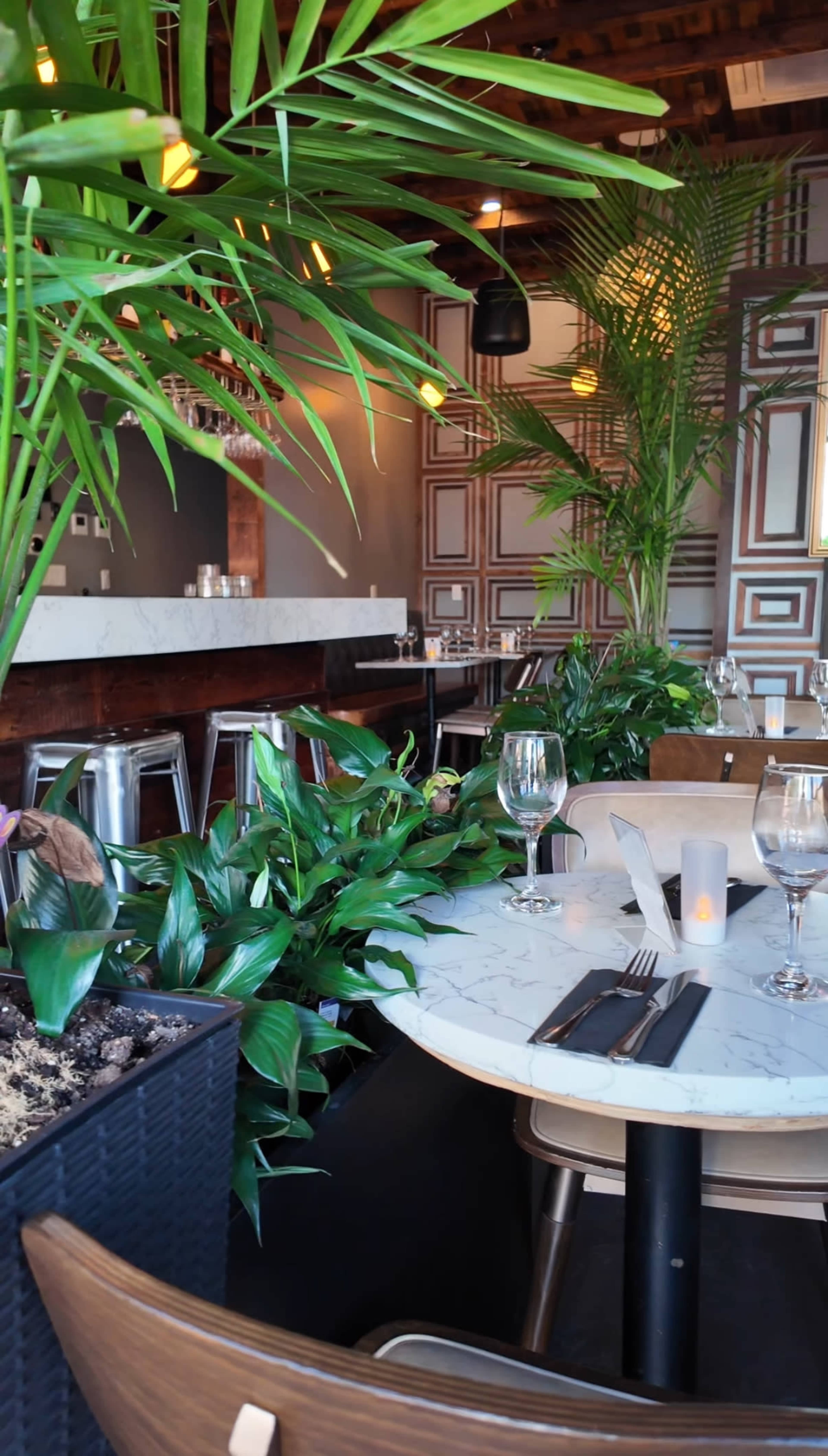 The image shows an indoor dining area of a restaurant featuring a marble table, glassware, and decorative plants amid a modern decor.