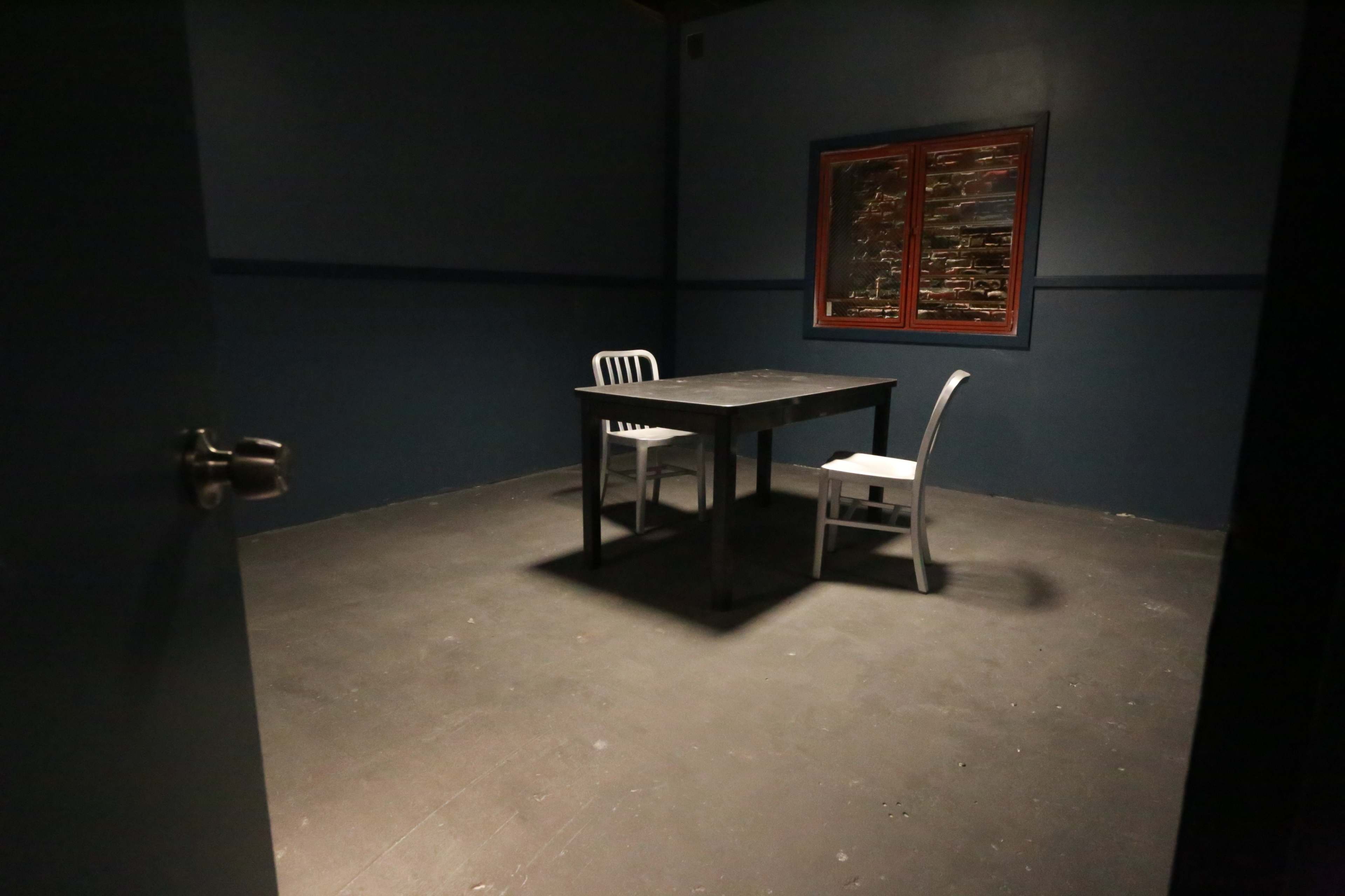 A sparsely furnished room with a dark floor, two white chairs, and a table under a window with covered glass.