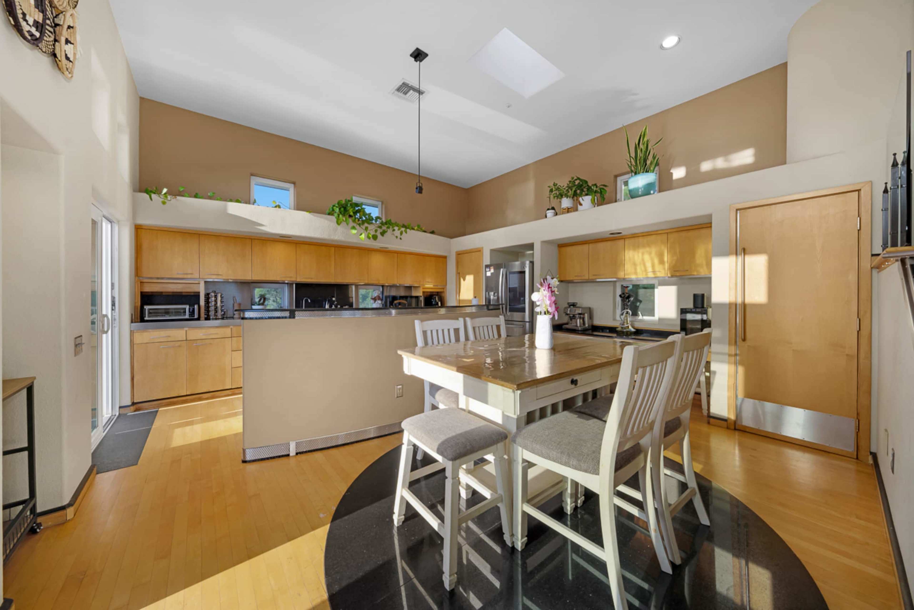 The image shows a modern kitchen with wooden cabinets, a central dining table, and large windows allowing natural light to enter the space.
