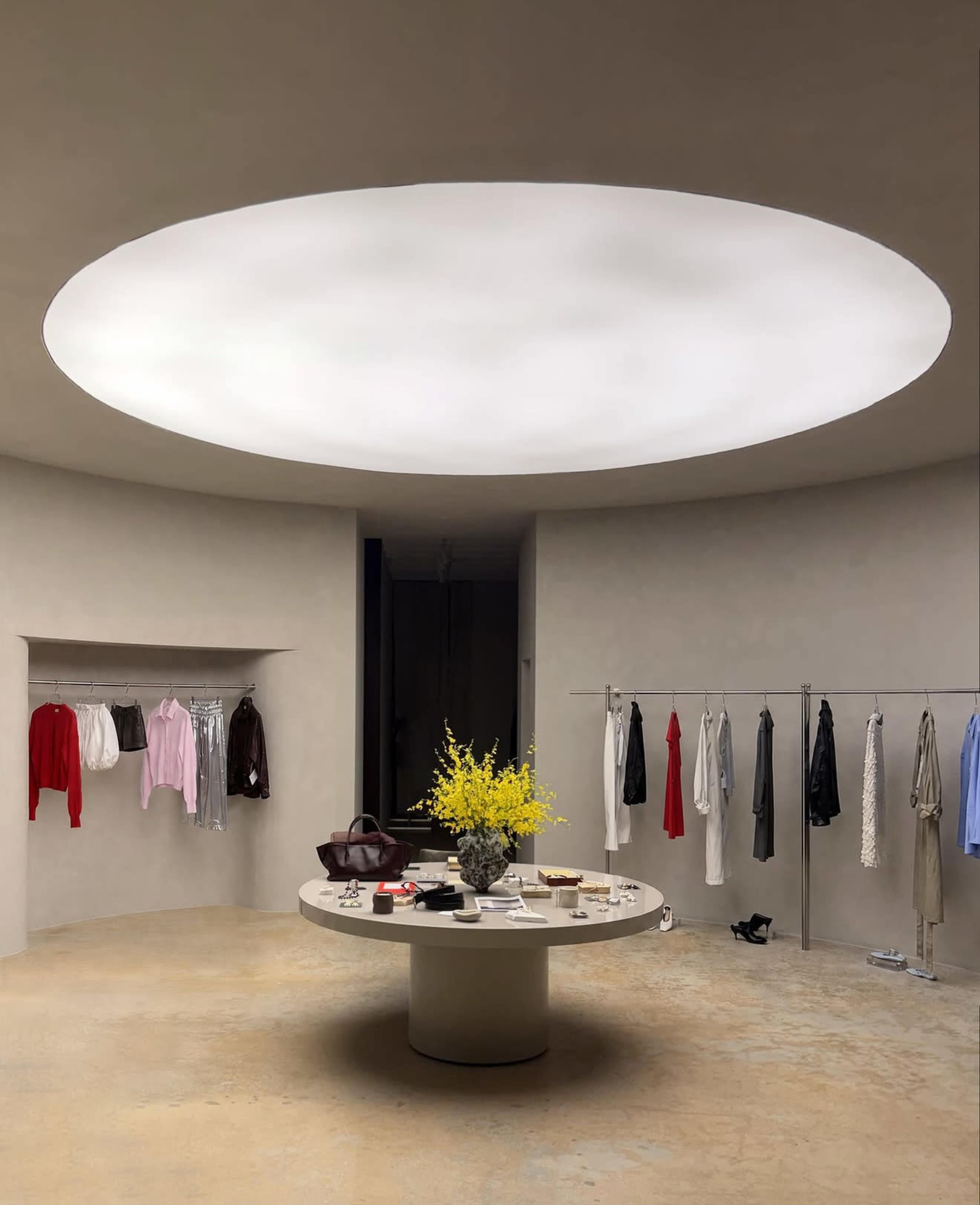The image depicts a modern retail space with a circular ceiling light, a central display table adorned with flowers and items, and clothing racks on two sides showcasing various garments.