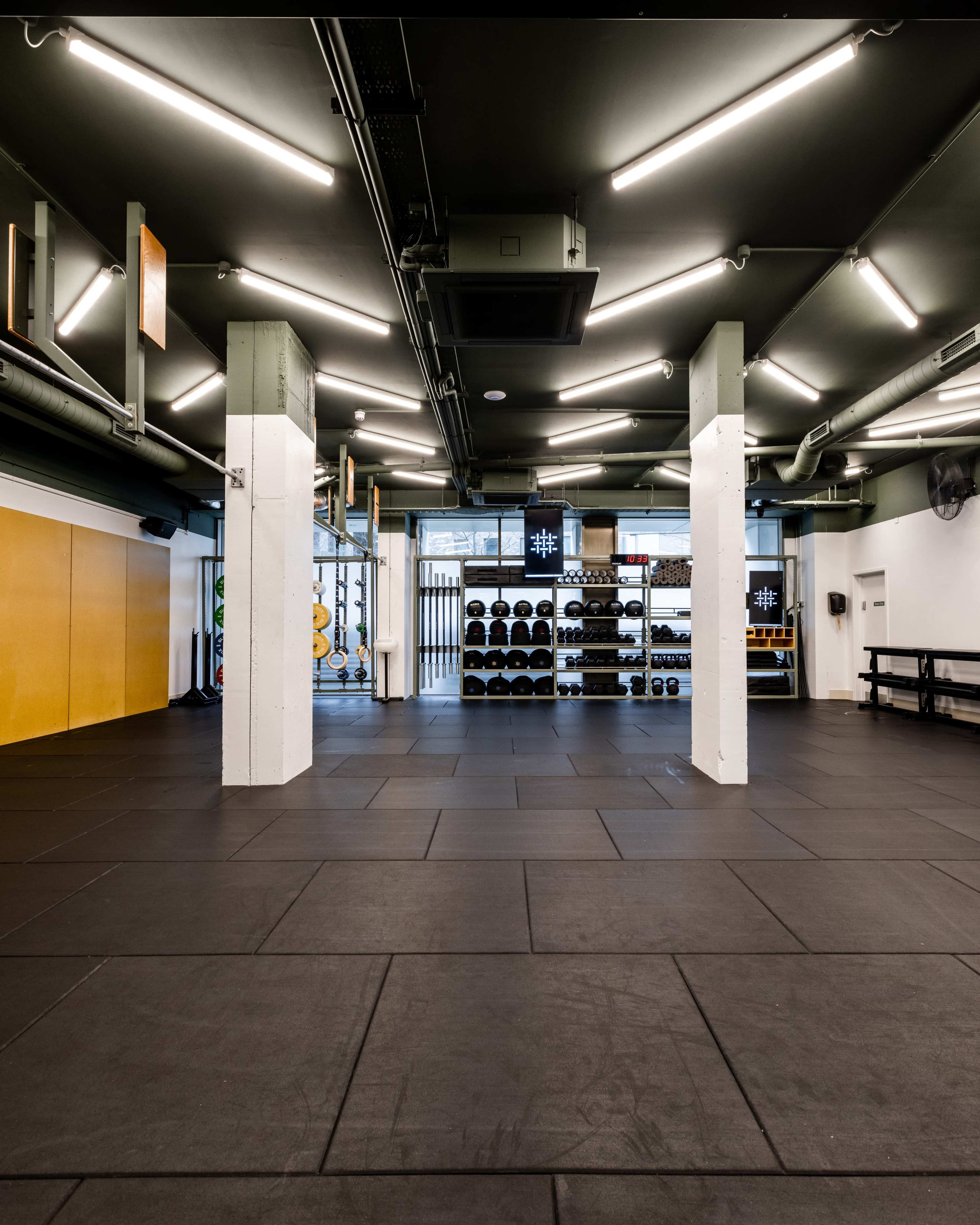 A spacious gym interior featuring rubber flooring, bright overhead lights, and a wall-mounted rack filled with dumbbells.