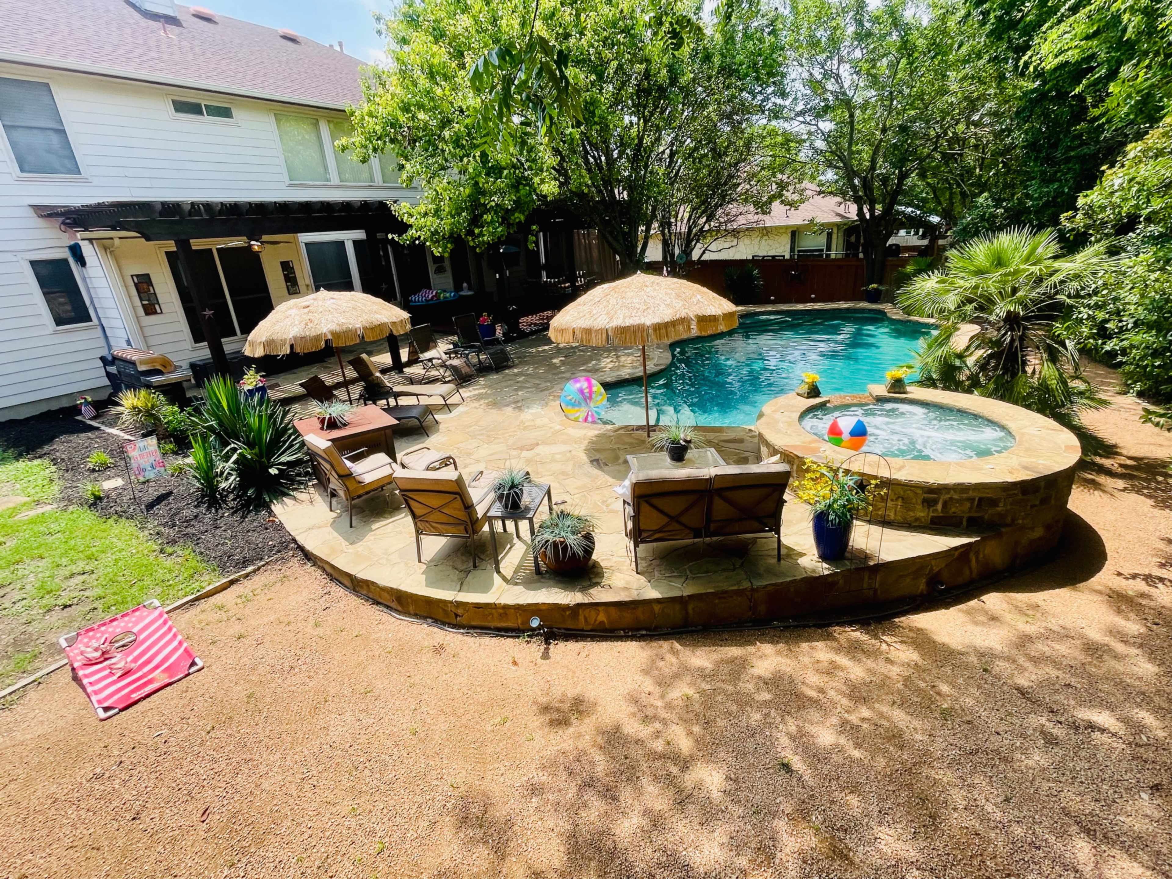 The image shows a backyard pool area with seating, umbrellas, and colorful pool floats surrounded by lush greenery.