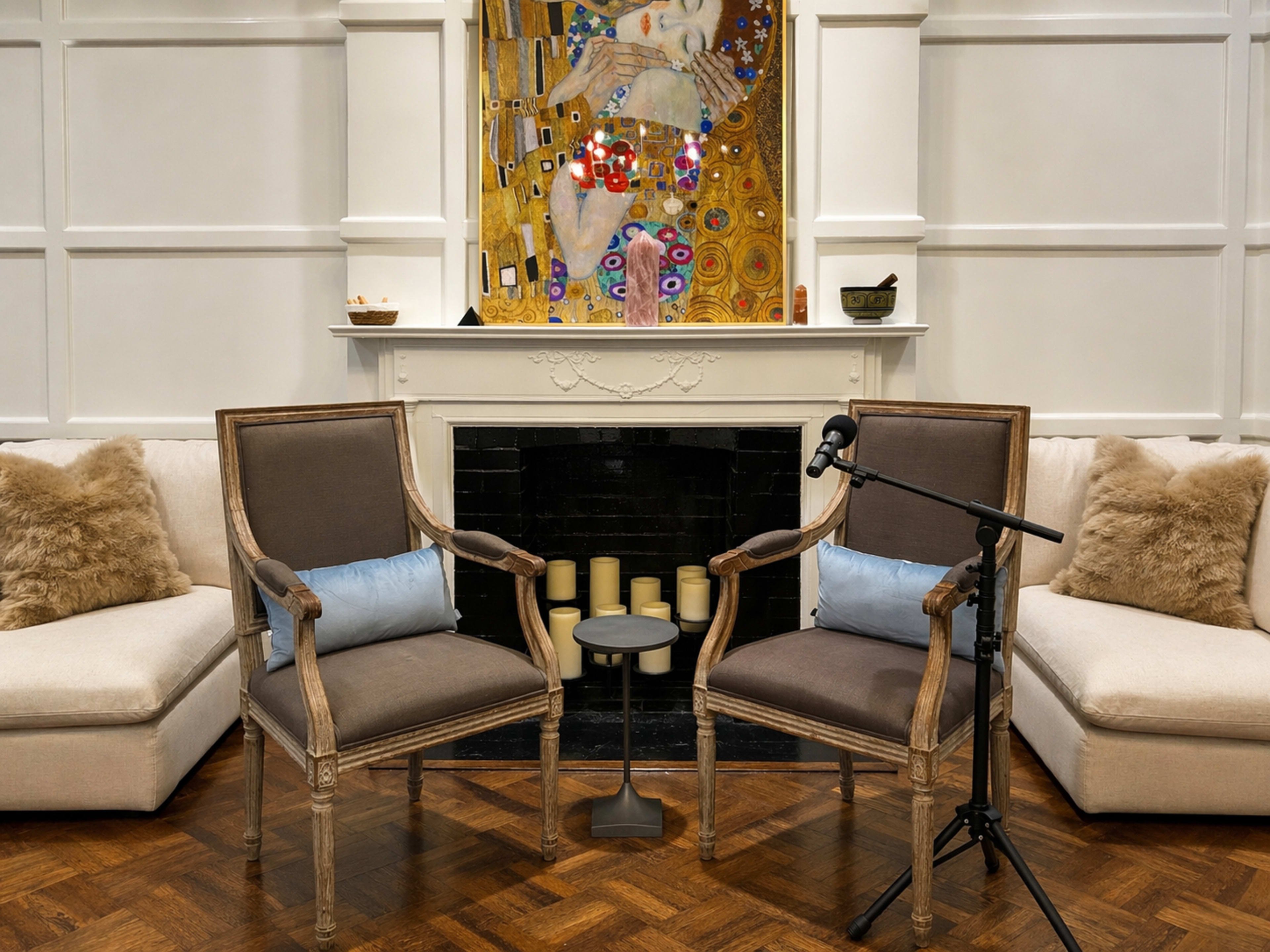 The image shows a cozy living room setup with two upholstered chairs facing a small table and a microphone, positioned in front of a fireplace, and decorated with a colorful artwork above the mantle.