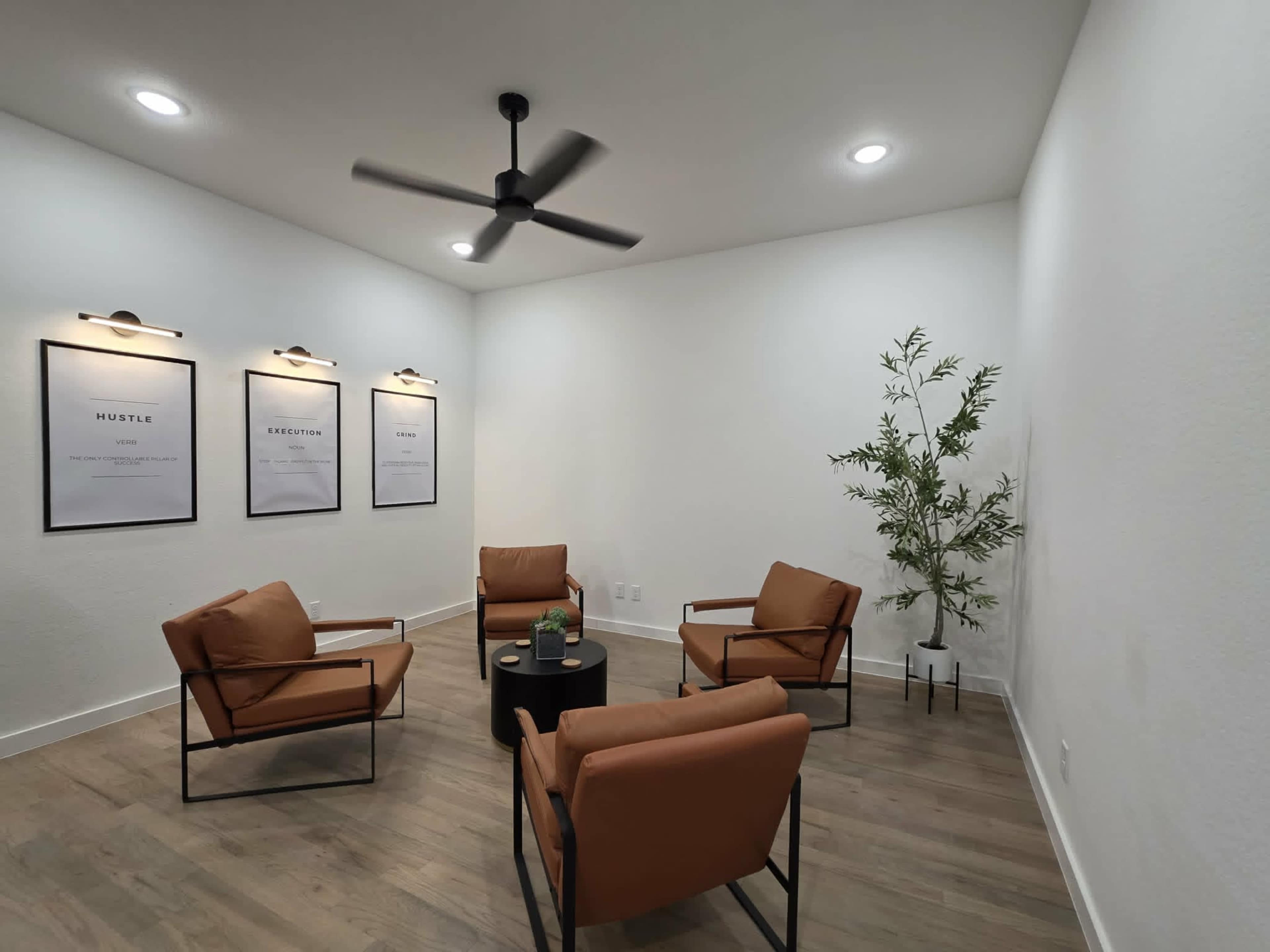 The image shows a minimalistic room with four brown chairs arranged around a small black table, featuring three framed posters on the wall and a plant in one corner.