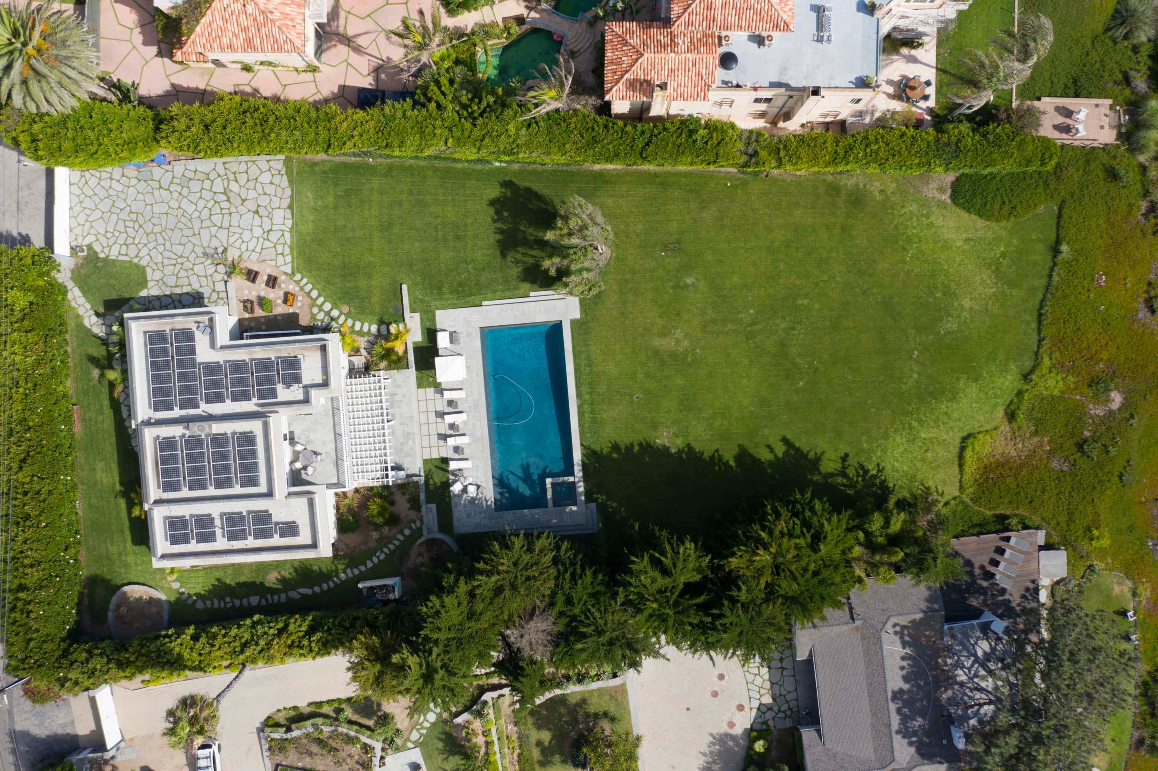 An aerial view shows a modern house with a swimming pool and solar panels, surrounded by a large green lawn and landscaped gardens.