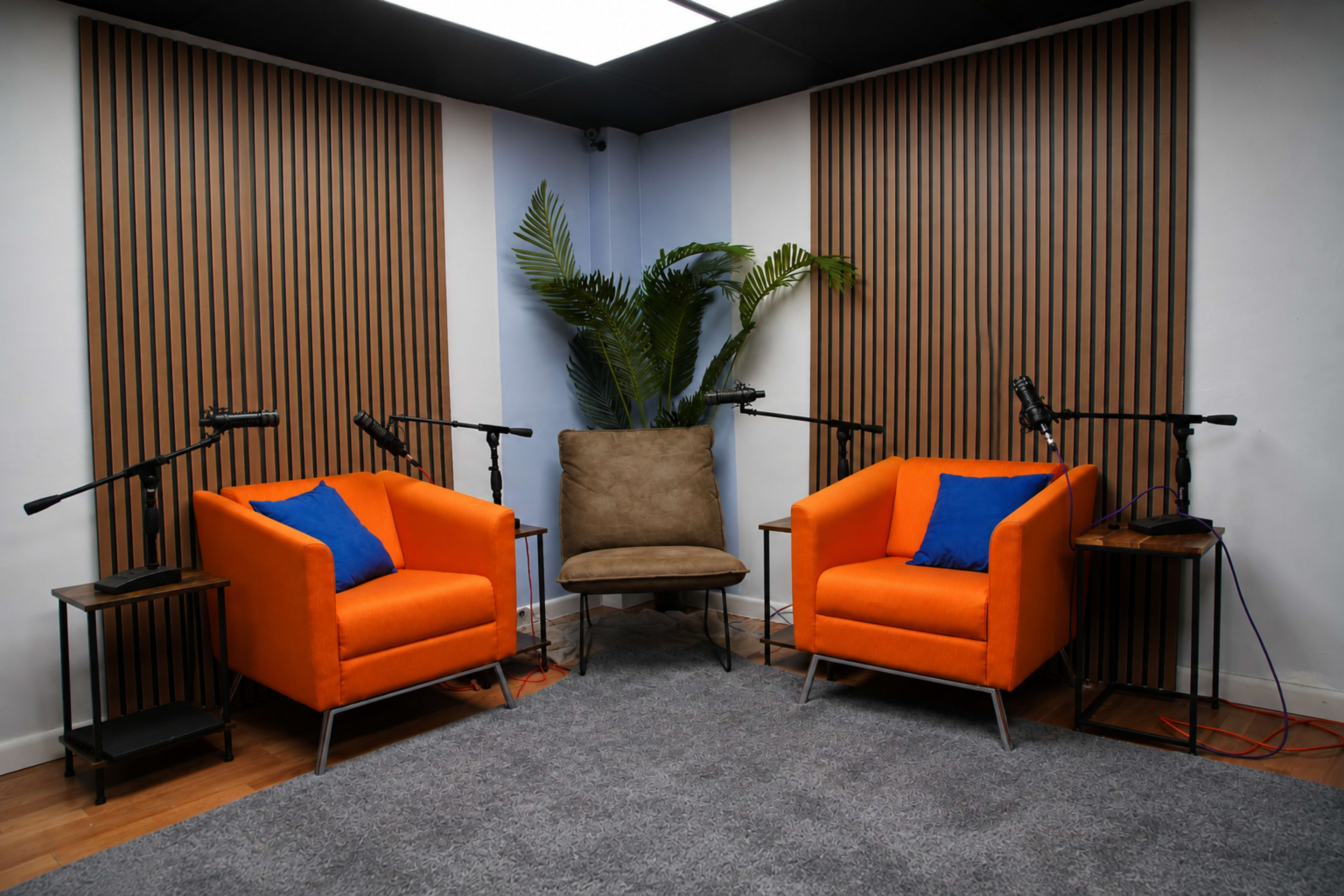 The image shows a modern podcast studio featuring two orange armchairs, a brown chair, and wooden paneling on the walls, with microphones positioned on stands.