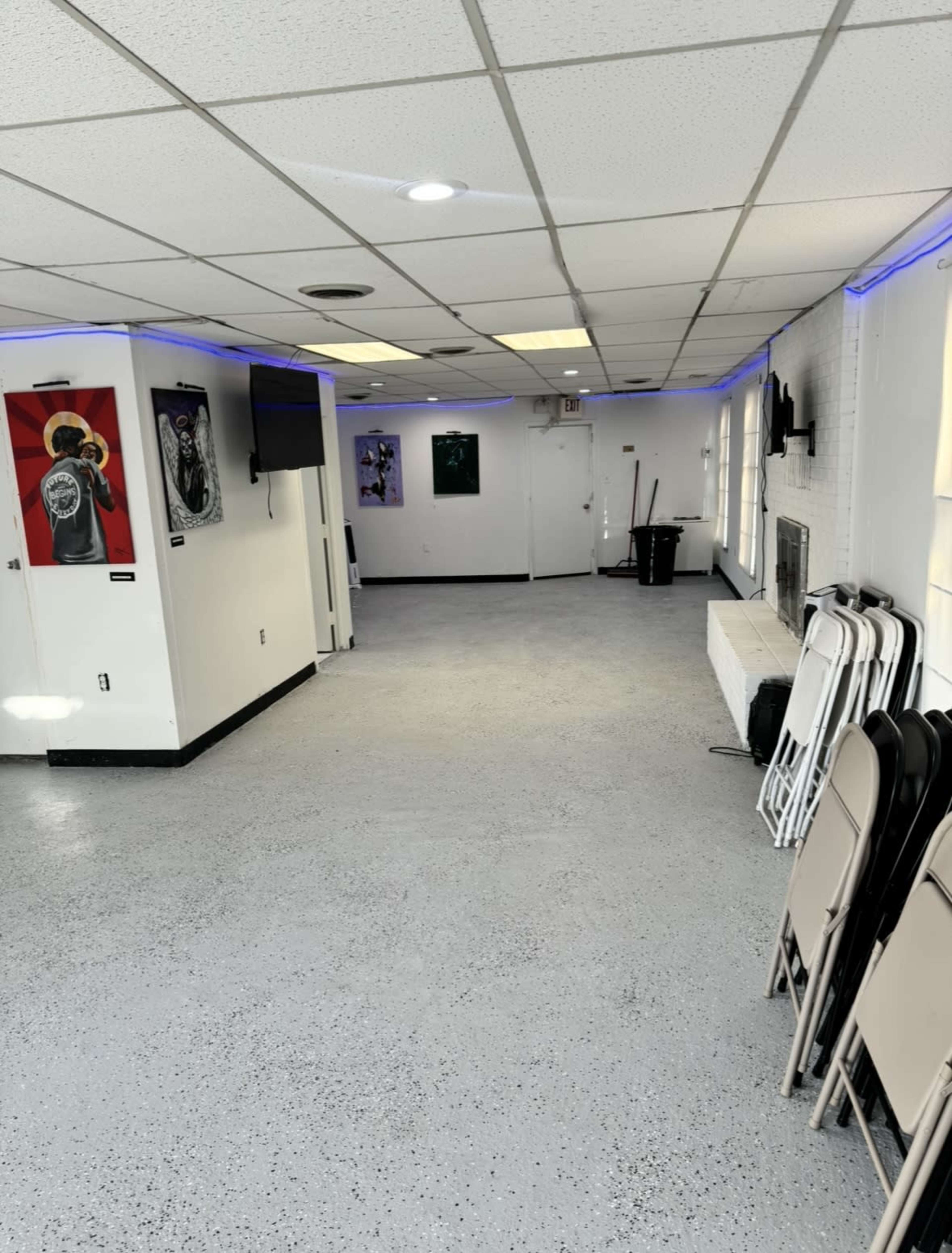 The image shows a spacious, well-lit room with a polished concrete floor, featuring several artworks on the walls and a set of folding chairs stacked against one side.