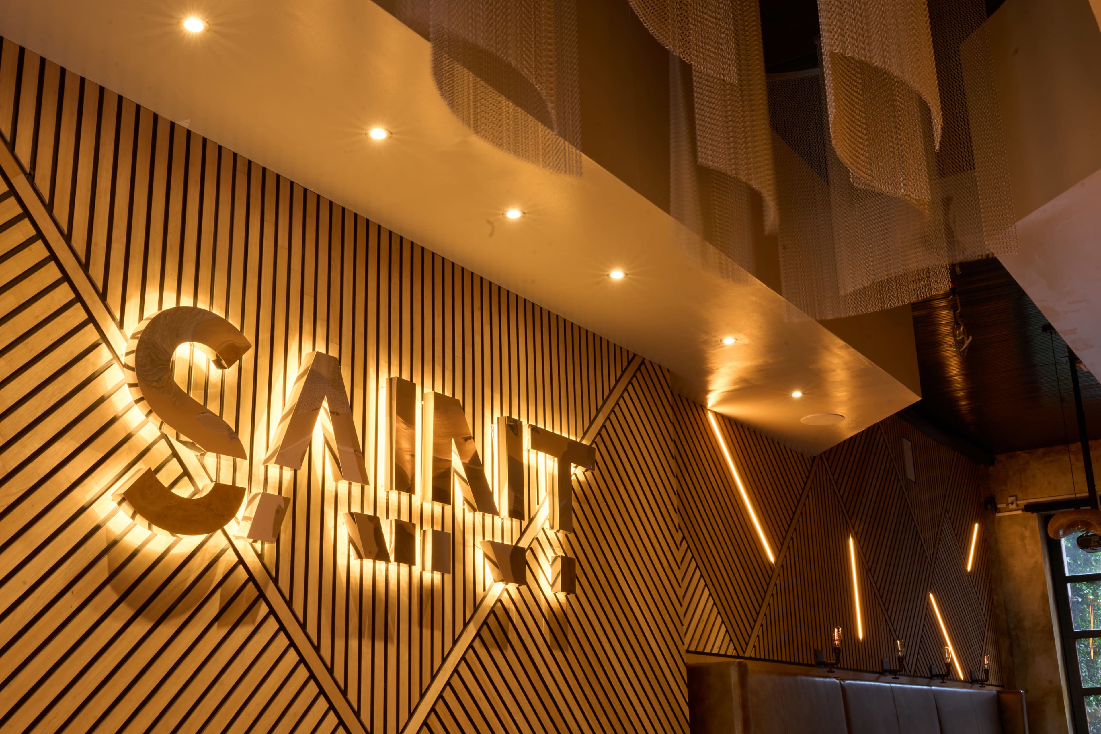The image features a modern interior of a restaurant or bar with wooden walls, illuminated text that reads "SAINT," and decorative lighting elements.