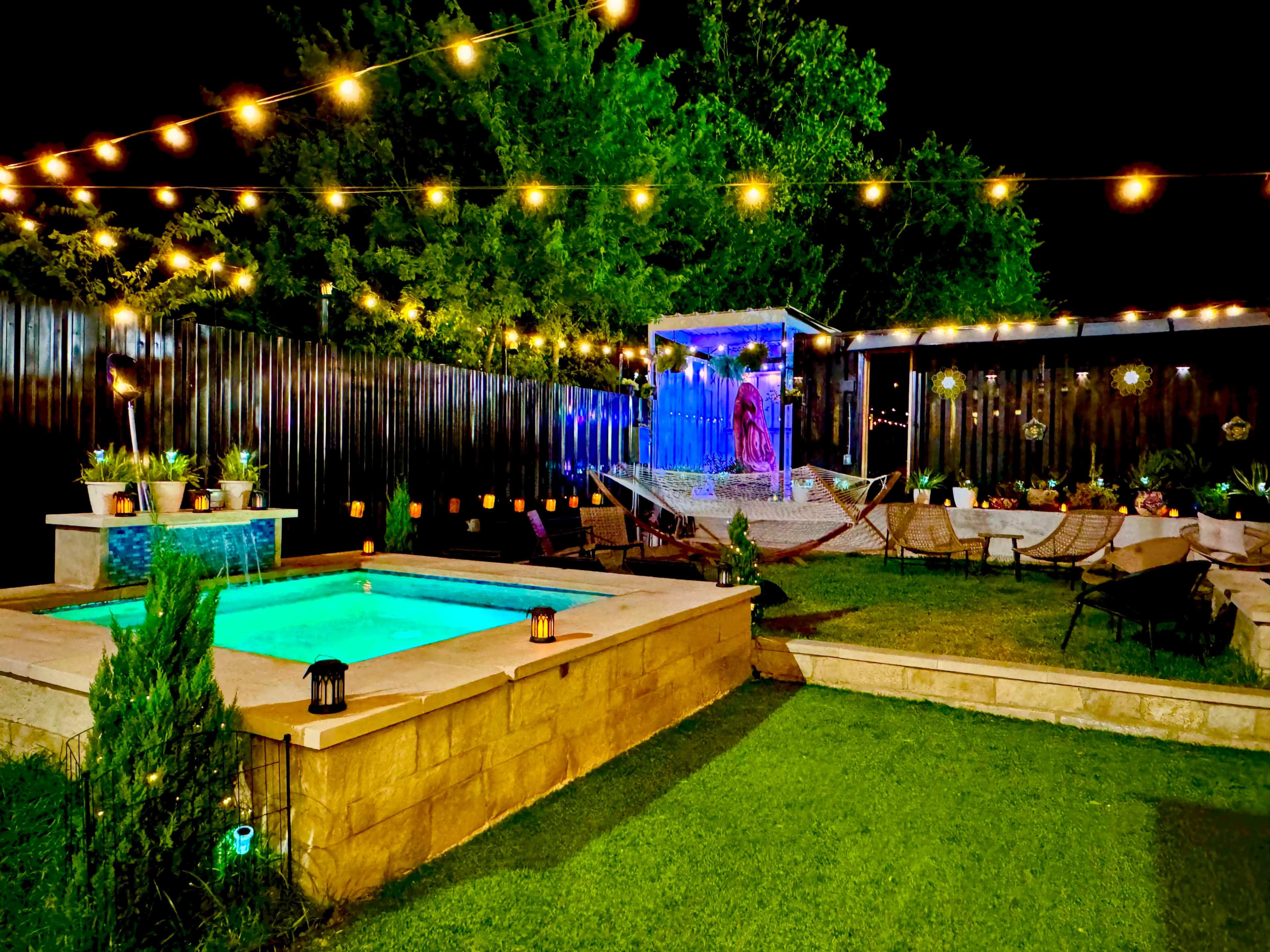 The image depicts a well-lit outdoor patio with a glowing turquoise pool, surrounded by greenery and decorative string lights.