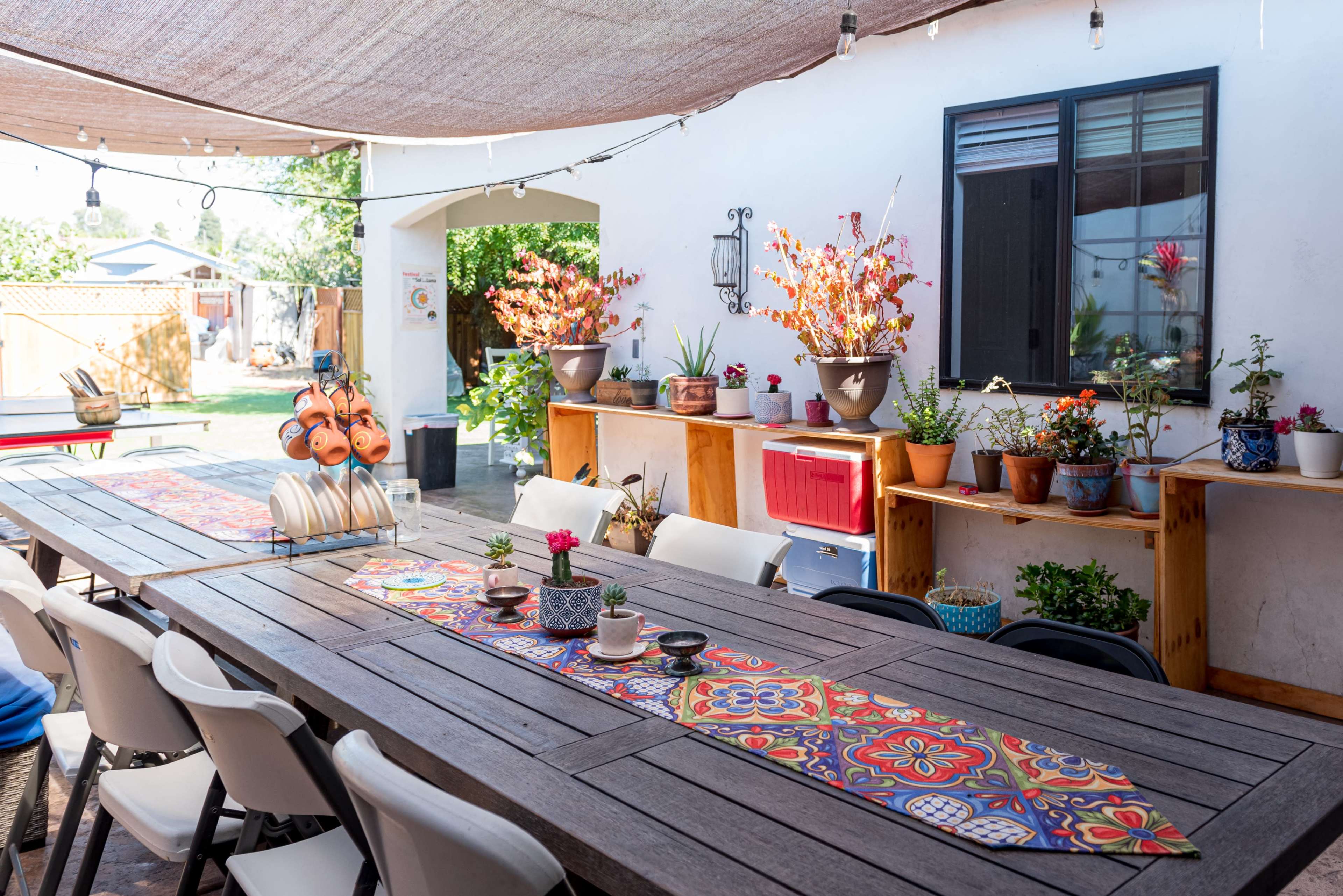 The image shows an outdoor patio with a long wooden table, adorned with colorful table runners and potted plants, under a shaded area with string lights.