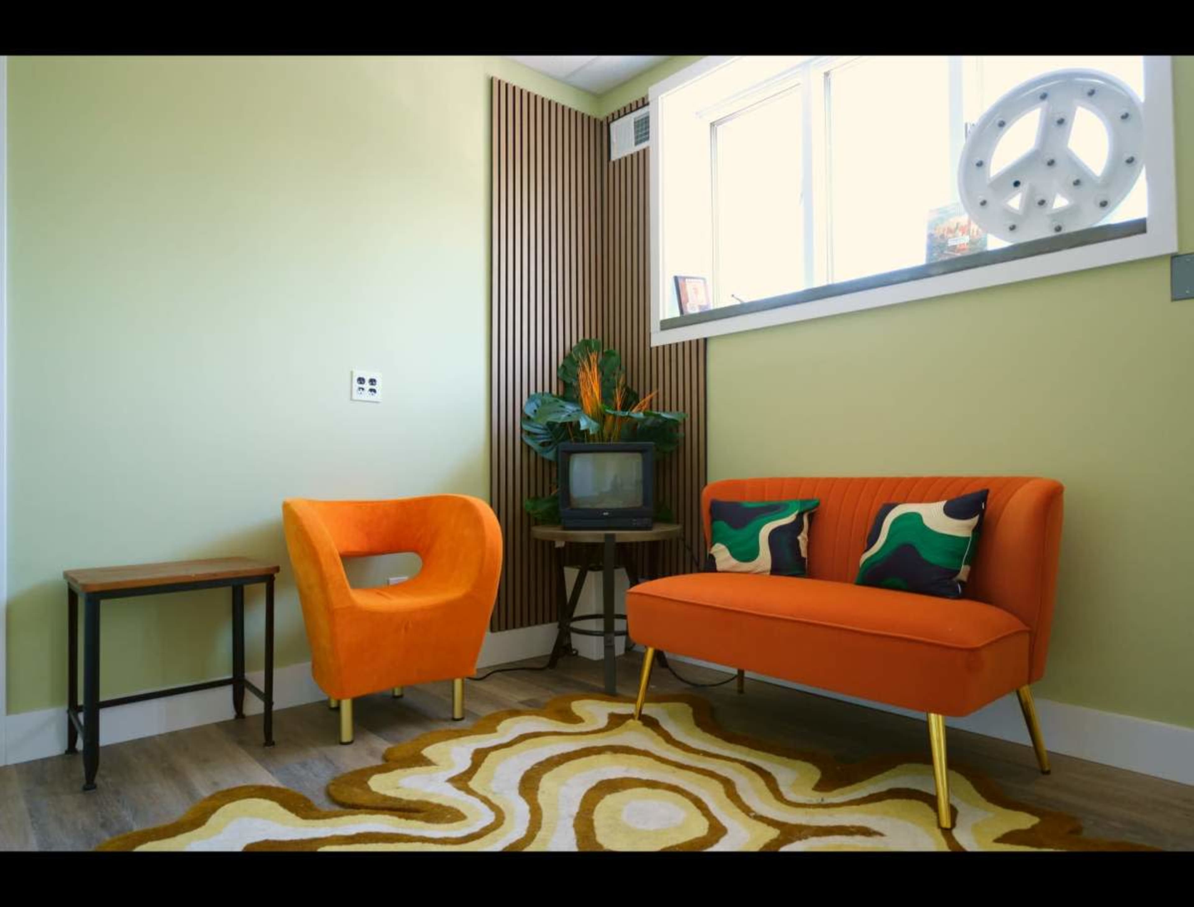 The image shows a brightly colored retro-inspired living space featuring an orange sofa, an orange chair, and a small wooden table on a wavy patterned rug.