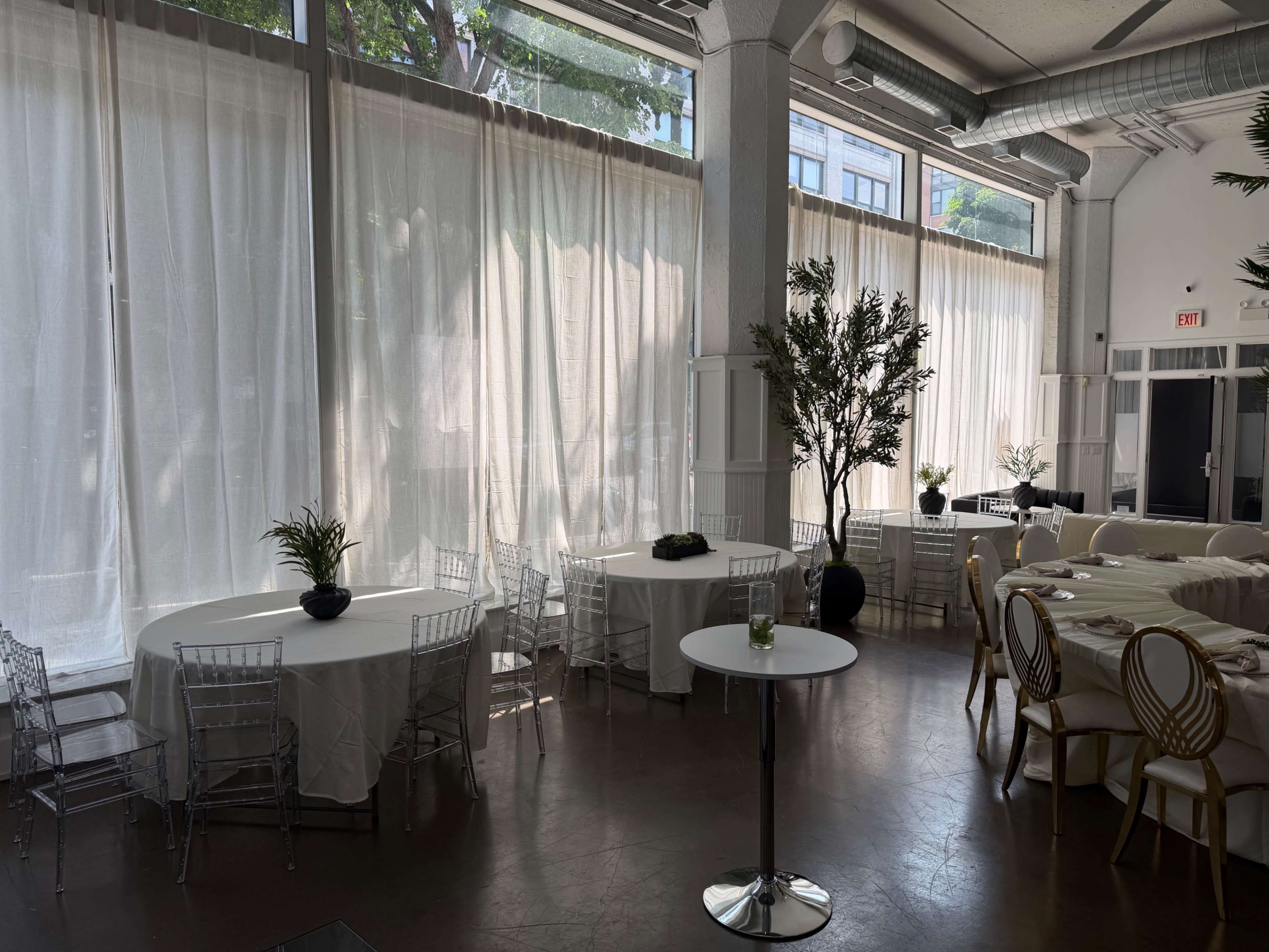 The image shows an indoor venue with large windows covered by sheer white curtains, featuring several tables arranged around the space.
