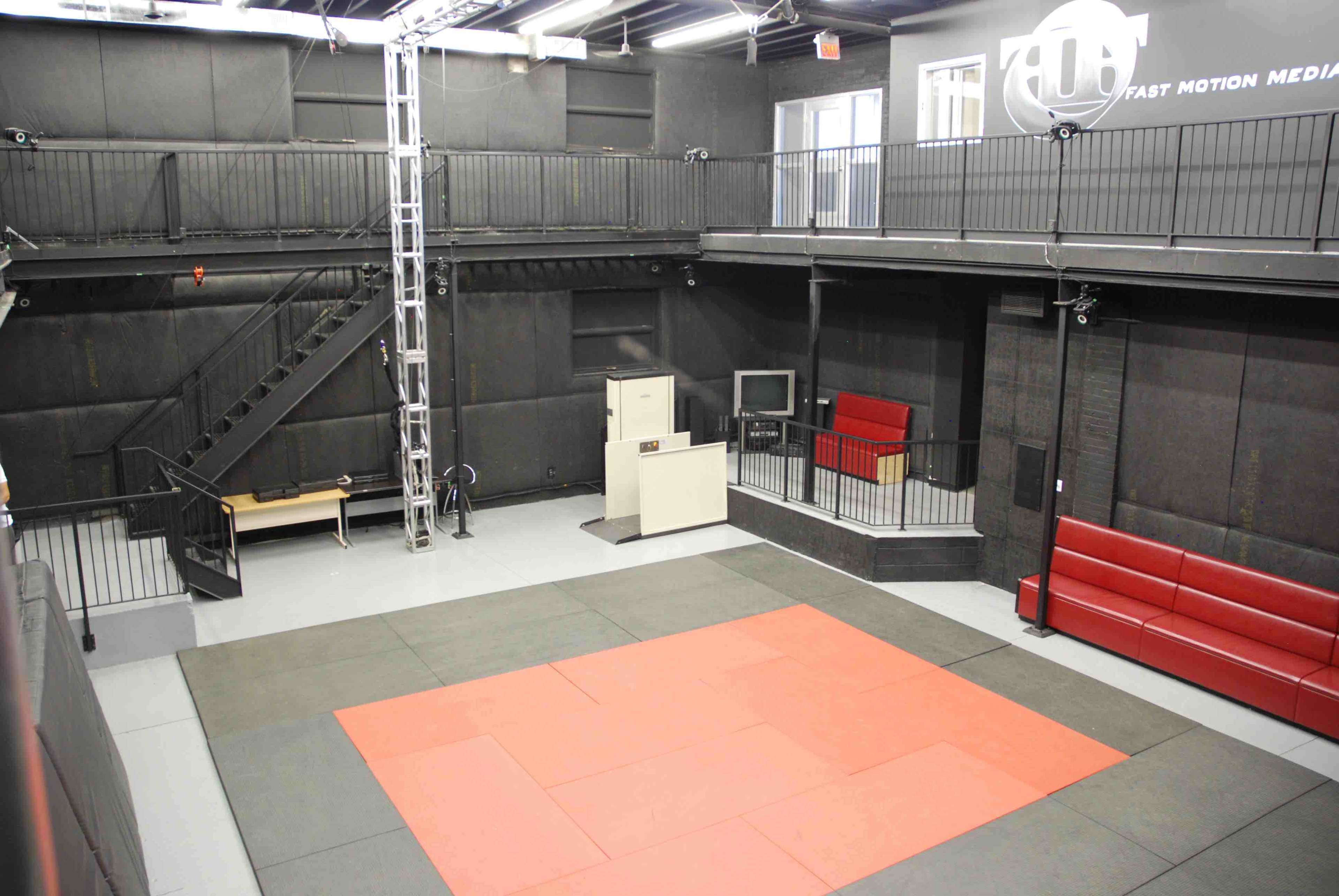 A spacious gym or studio with a padded training area, a staircase, and seating along the walls.