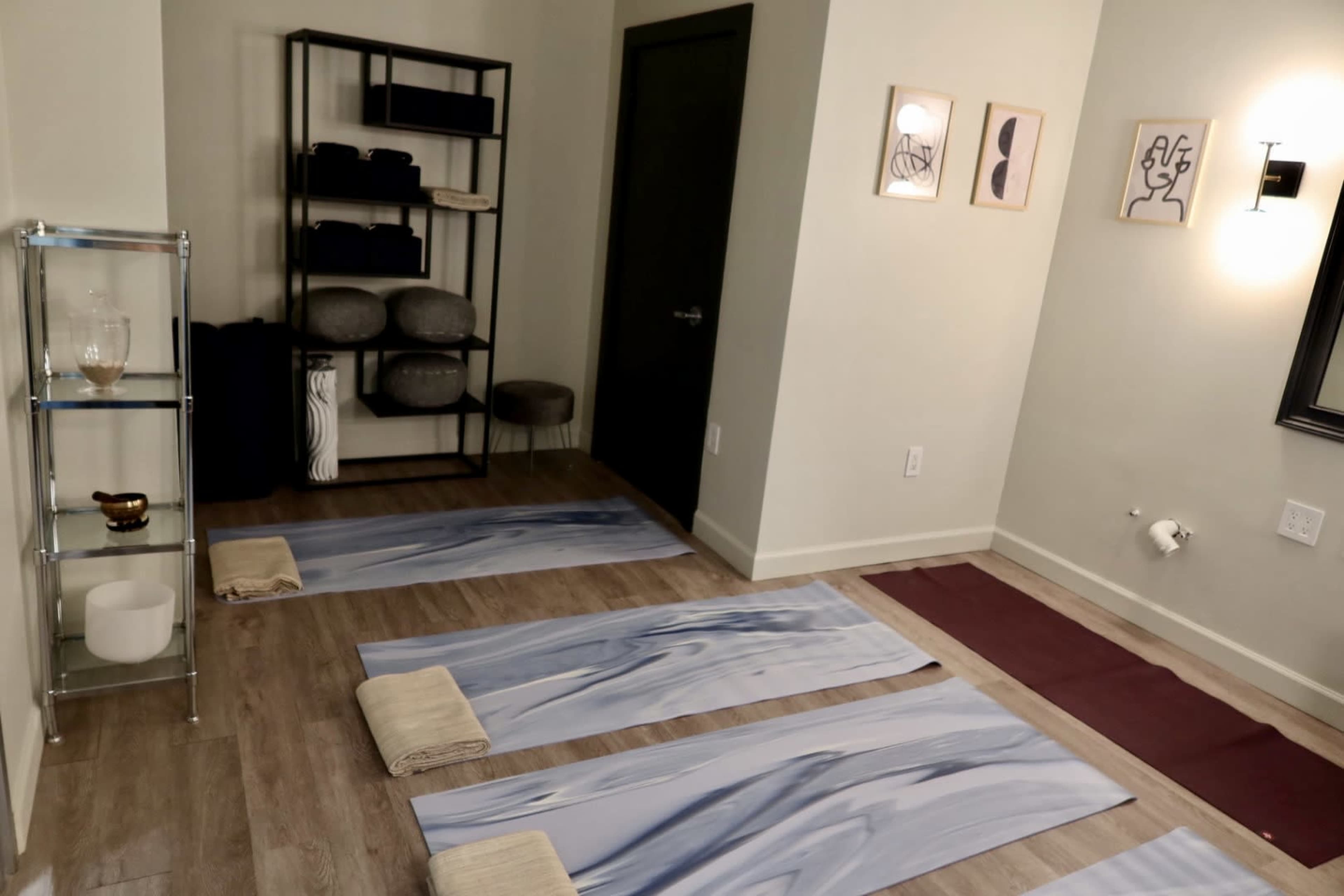 The image shows a serene indoor space with yoga mats laid out on a wooden floor, shelves with folded towels, and framed artwork on the walls.