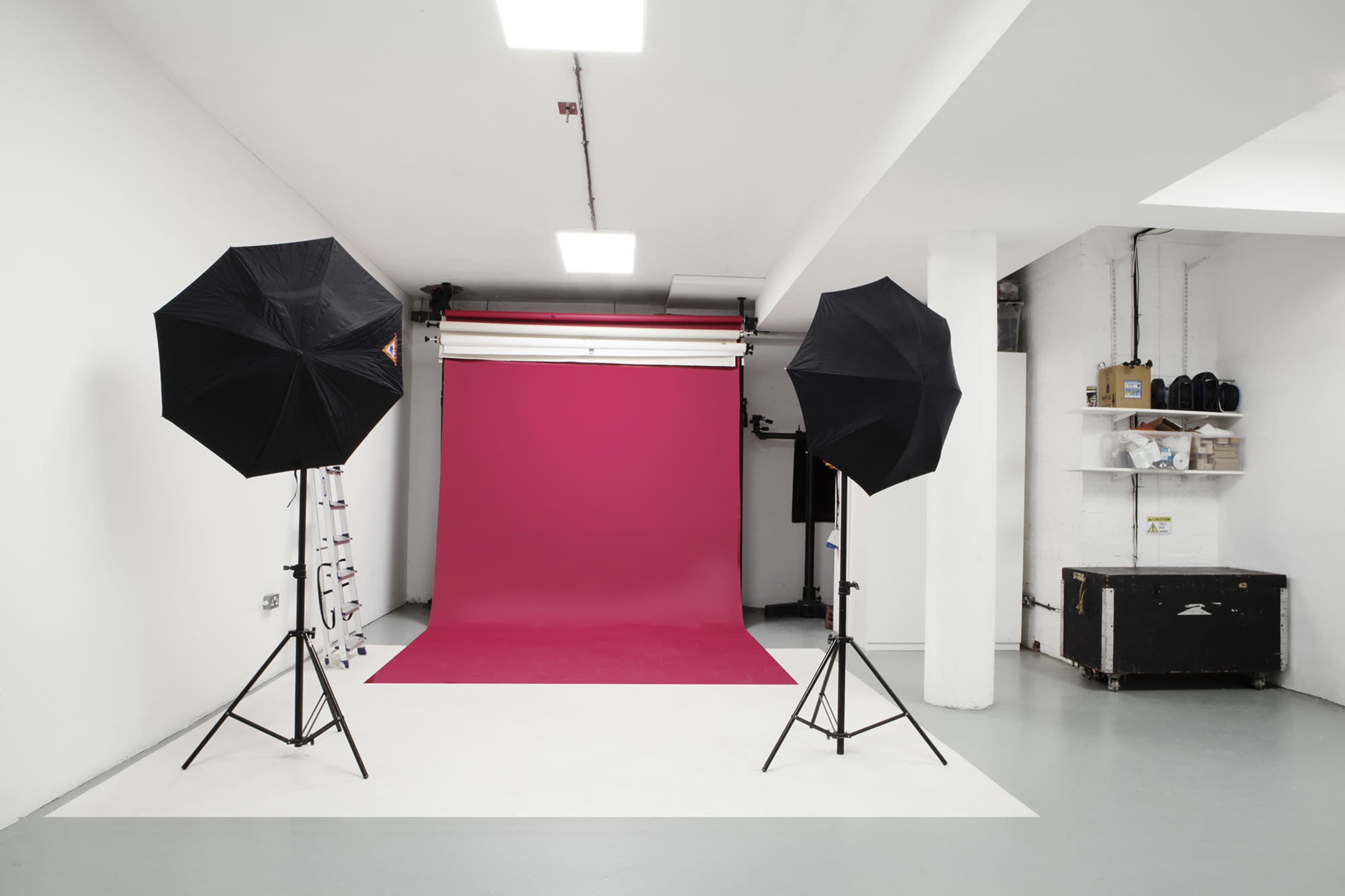 A photography studio with a pink backdrop, two black umbrella light stands, and a white floor.