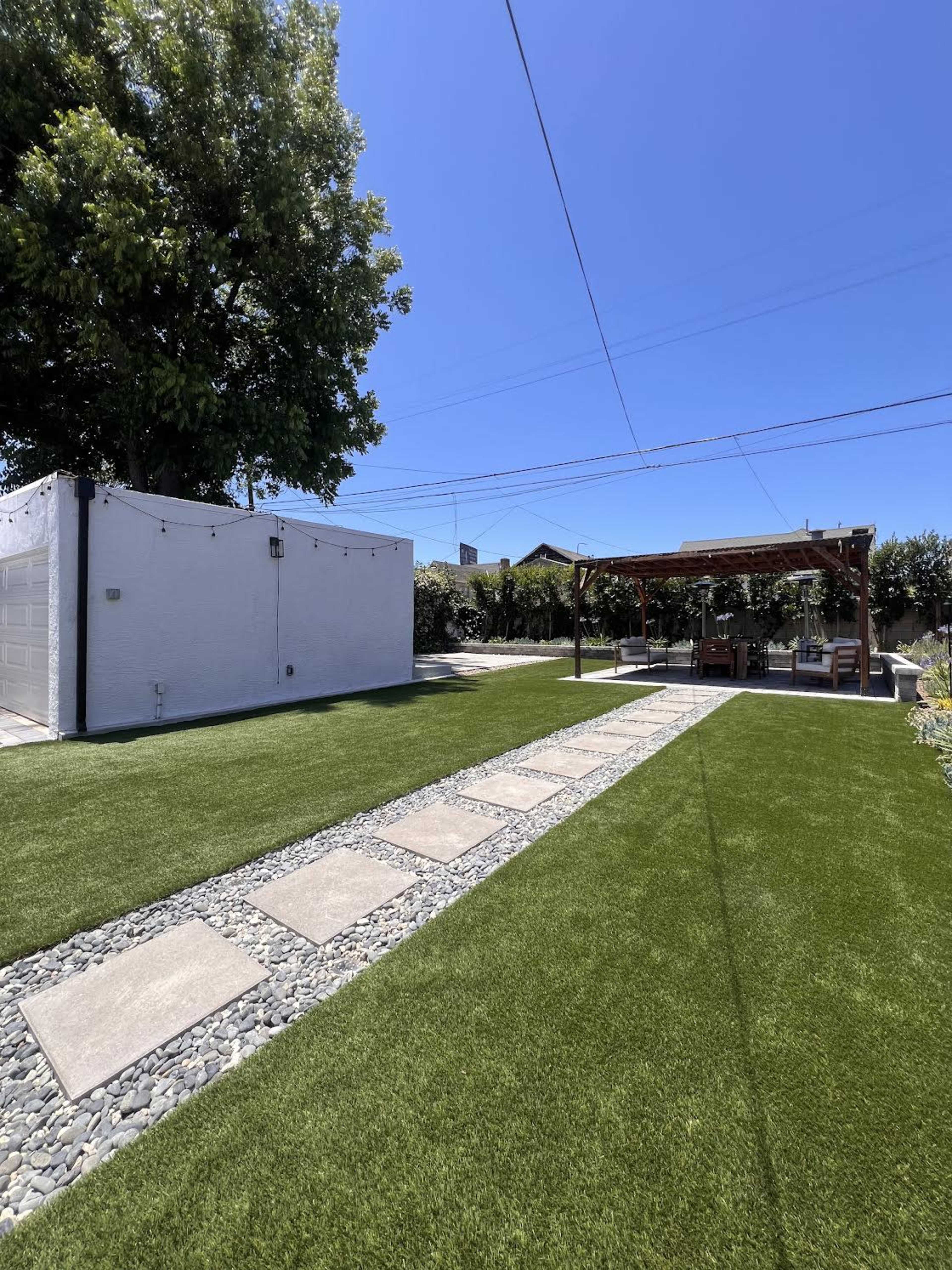 The image shows a landscaped backyard with artificial grass, stone pathways, and a wooden pergola providing shaded seating.
