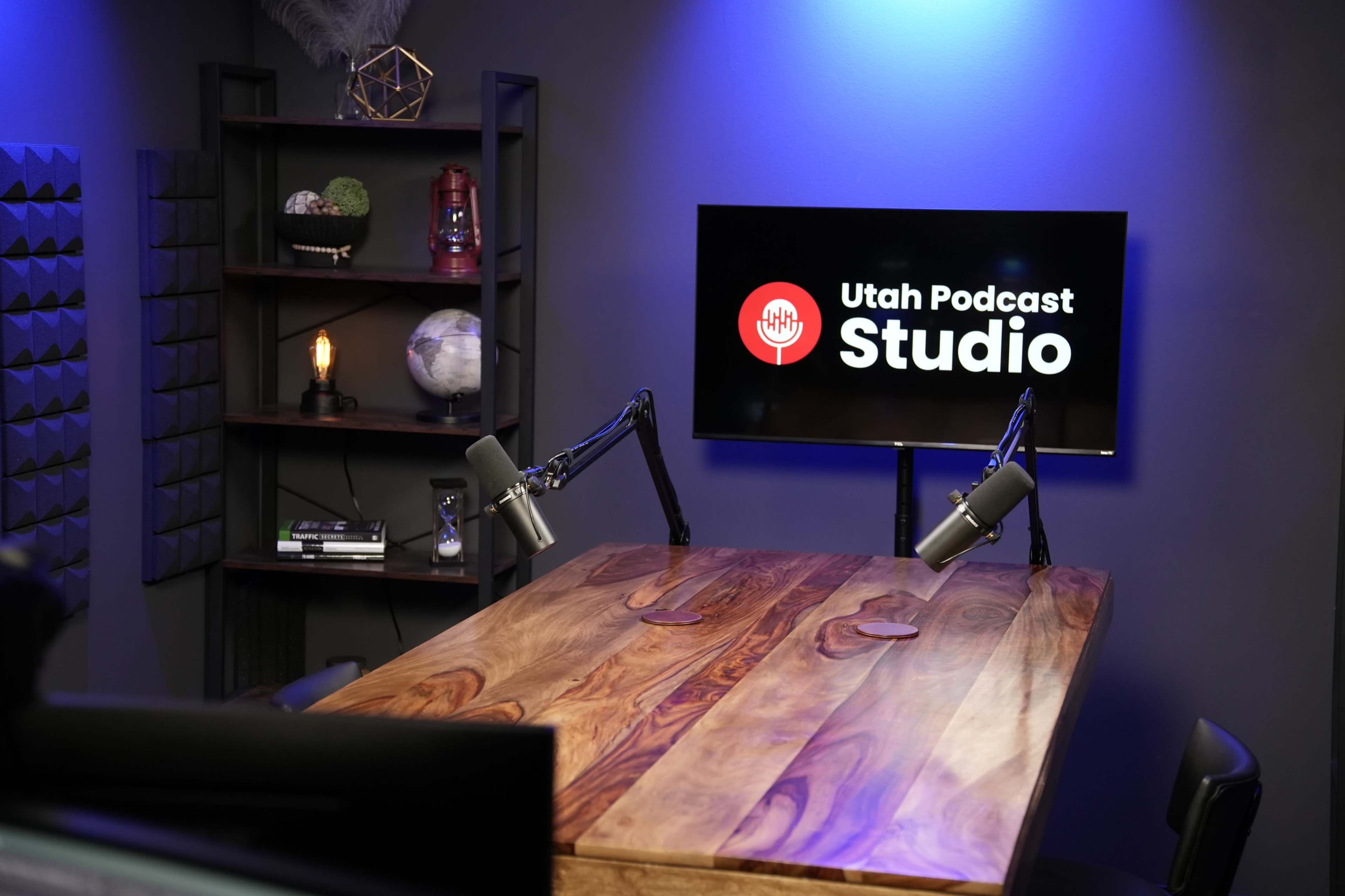 The image shows a podcast studio with a wooden table, two microphones on adjustable arms, and a screen displaying "Utah Podcast Studio" against a blue-lit wall.
