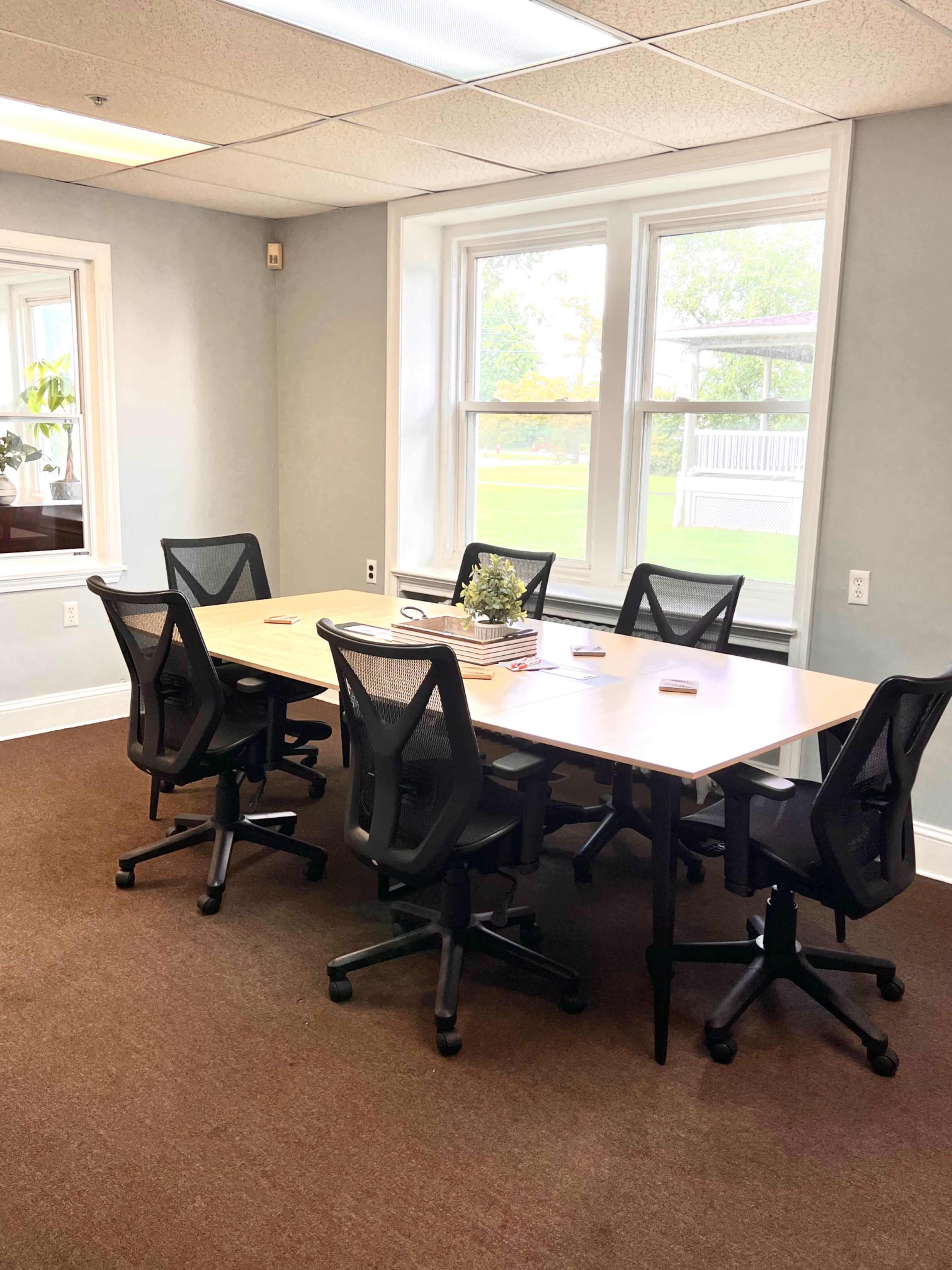 A modern conference room features a large table surrounded by six ergonomic office chairs, with windows providing natural light.