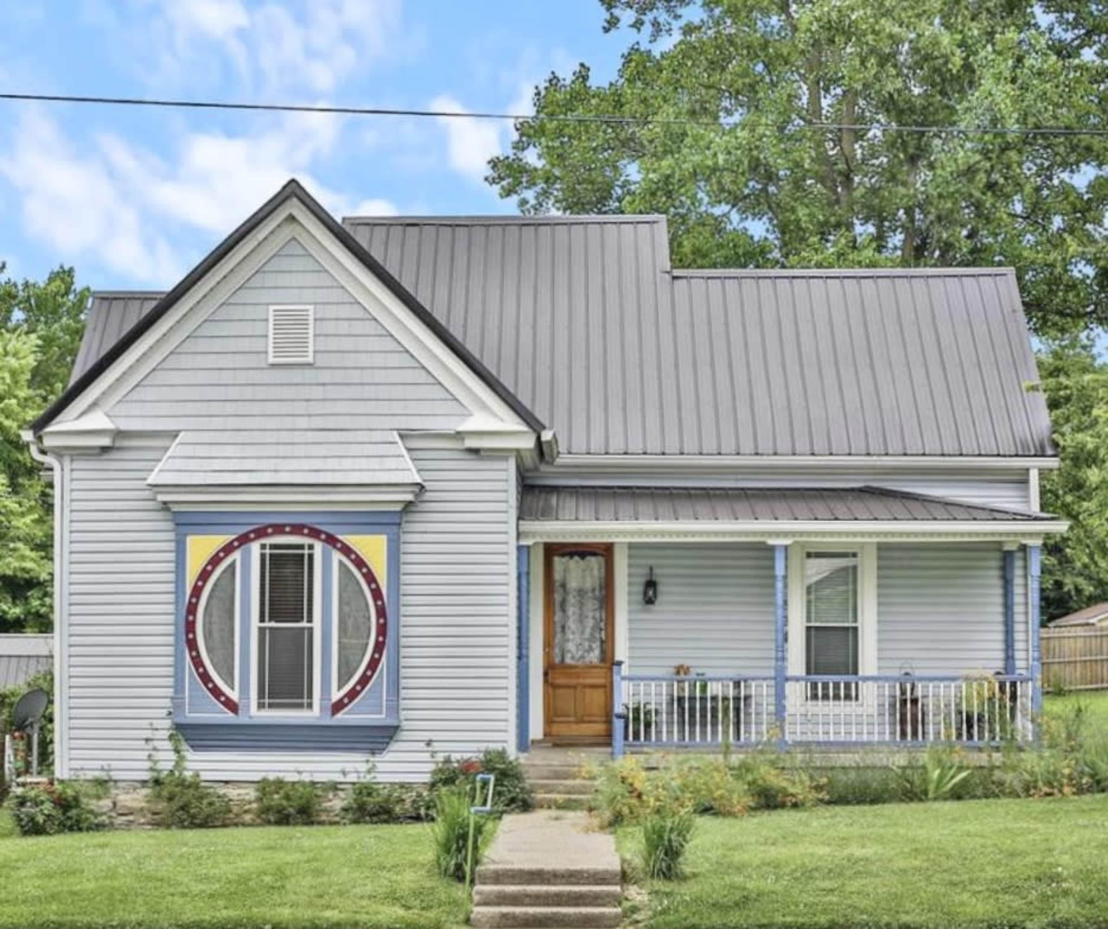 A light blue house with a metal roof, featuring a decorative painted circle on the front wall and a porch with white railings.