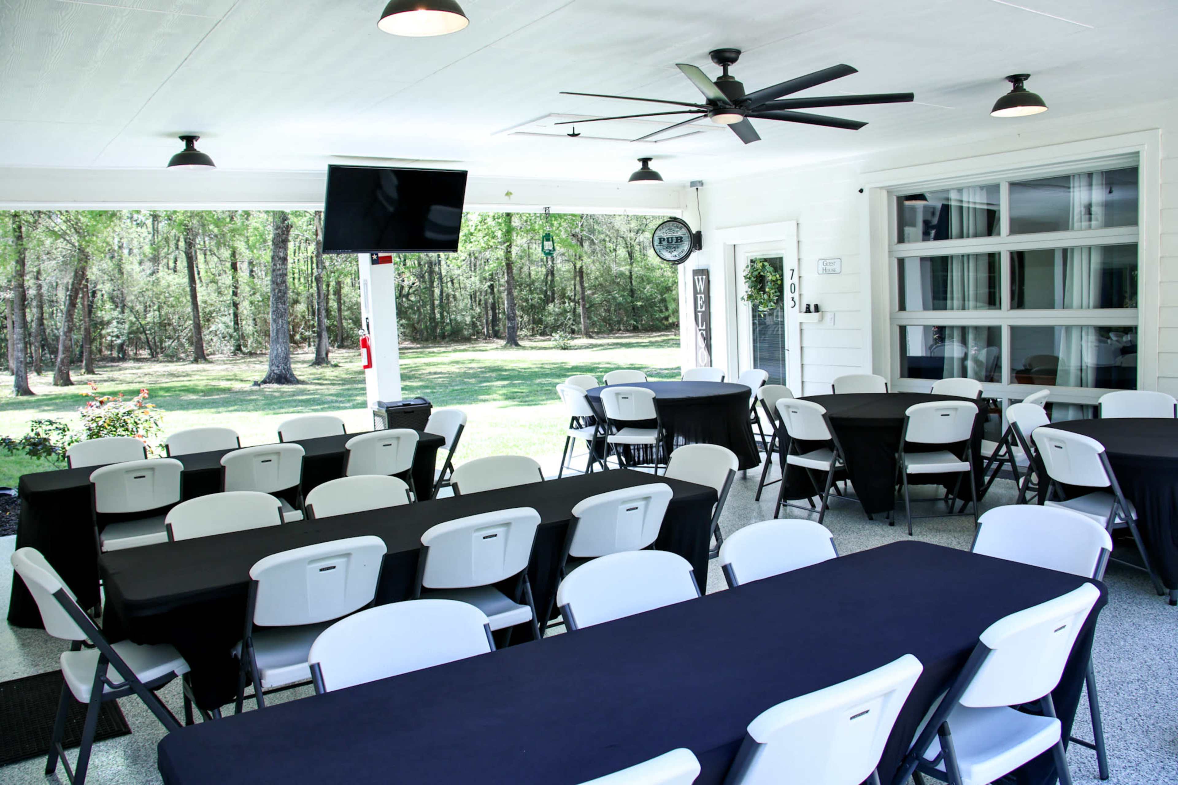 An outdoor seating area with several black and white tables and chairs arranged under a covered porch, flanked by large windows and a television.