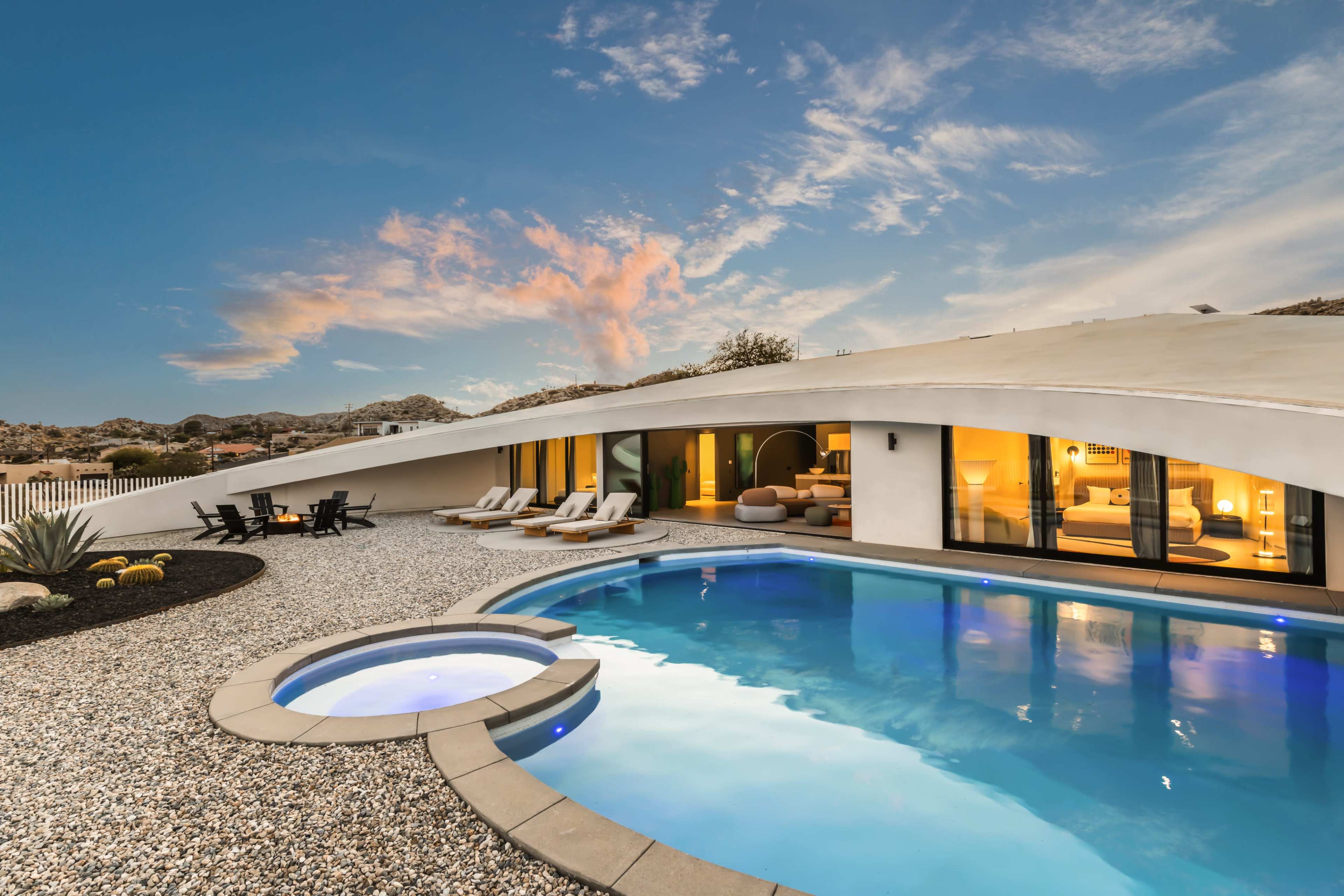 The image shows a modern home with an outdoor pool surrounded by gravel and lounge chairs, set against a backdrop of a colorful sky at sunset.