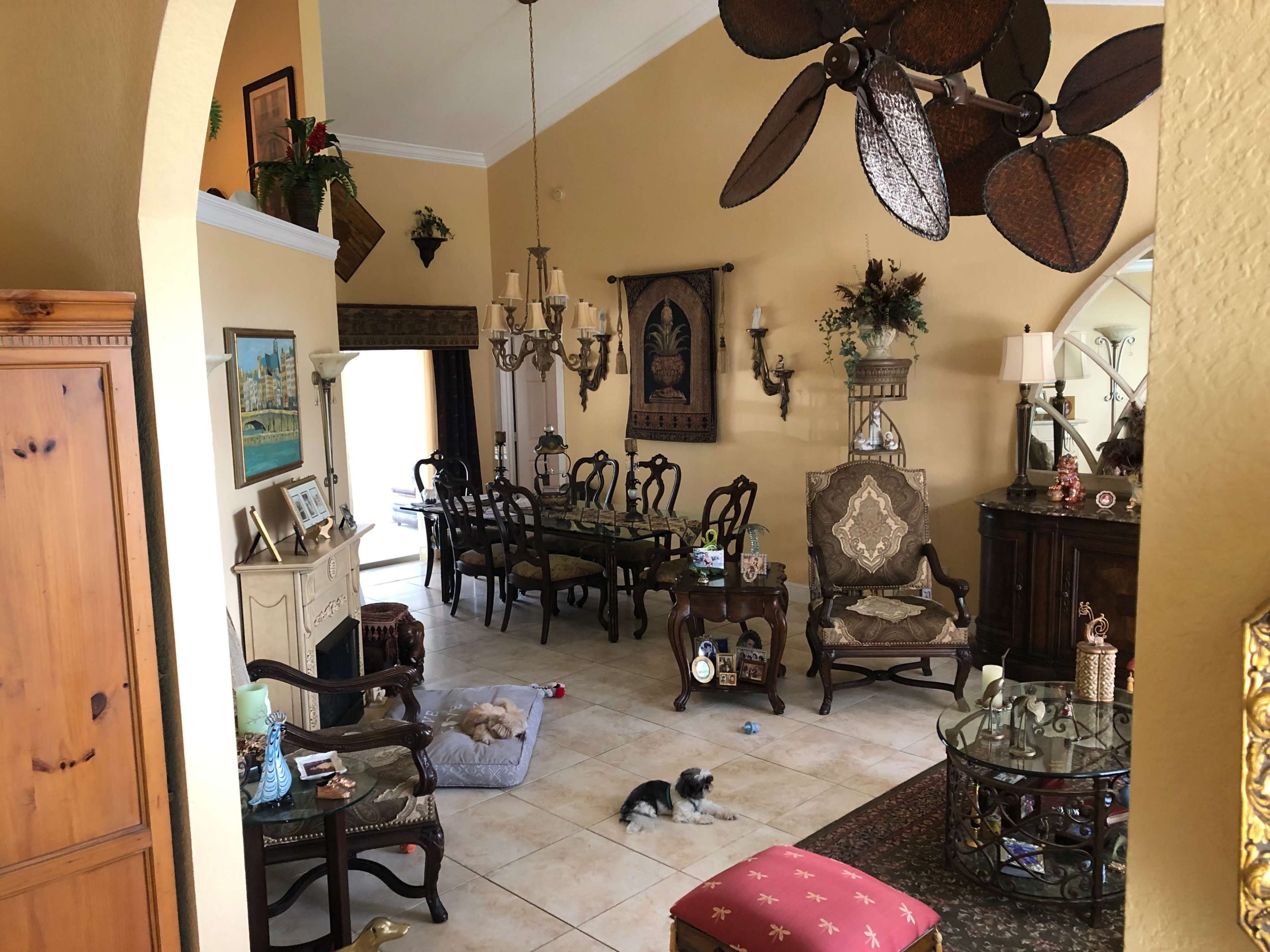 A spacious living room features a dining area with a large table, elegant furniture, and two small dogs resting on the tiled floor.