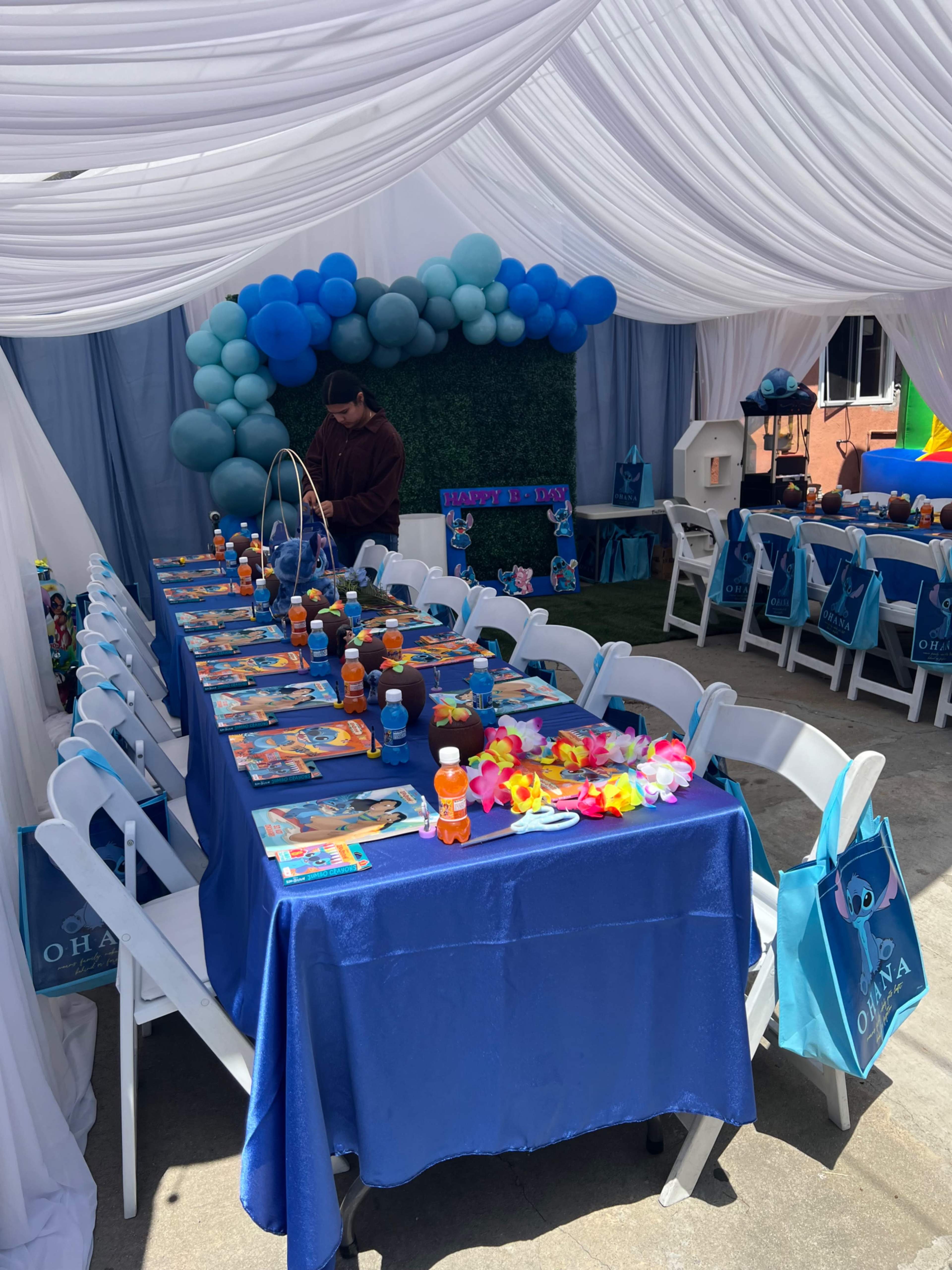 A decorated outdoor party space features a long table with blue tablecloths, colorful party favors, and balloons, surrounded by white drapery and chairs.