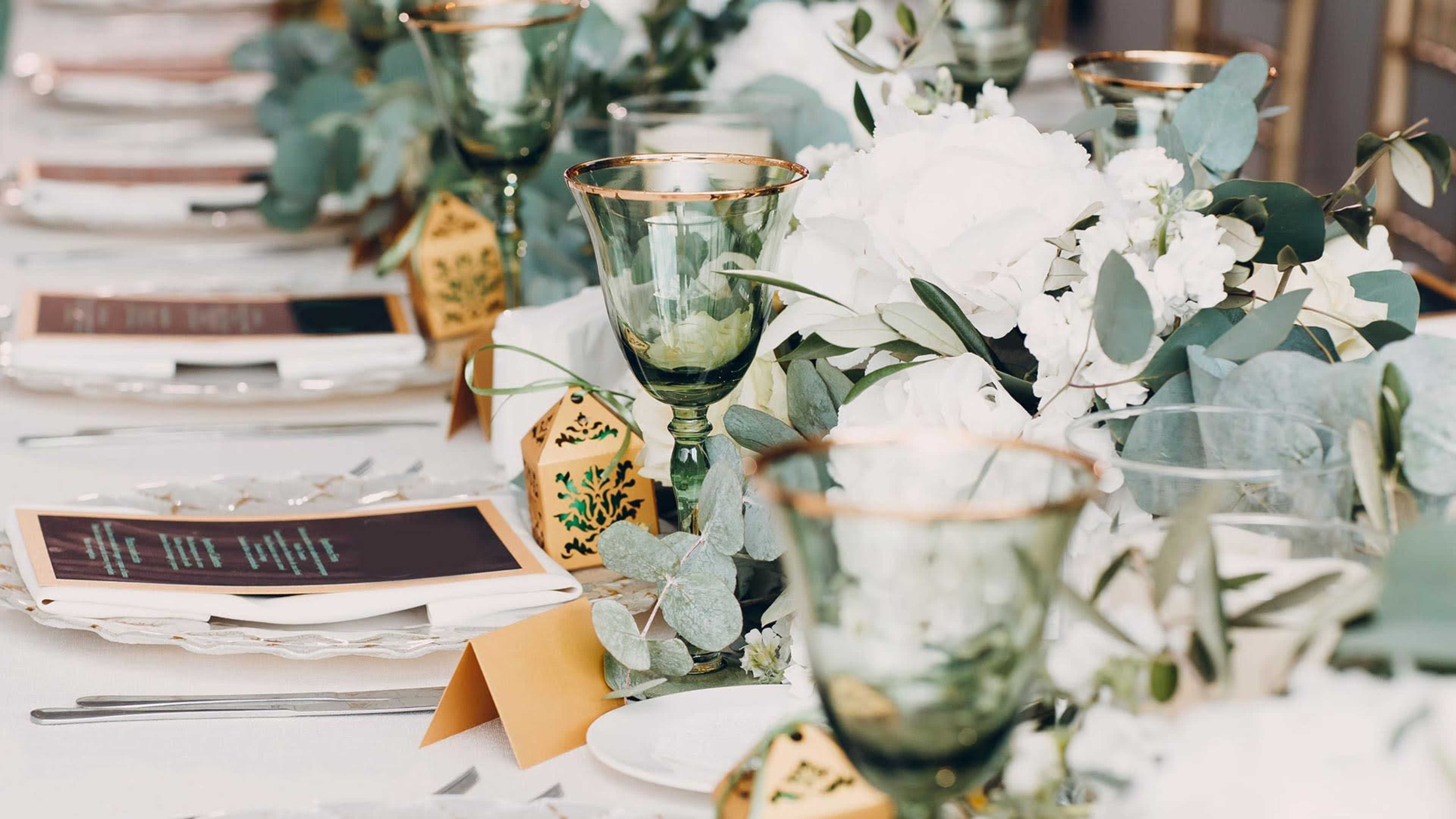 A long dining table is set with green glassware, white floral arrangements, and menus beside neatly arranged plates.