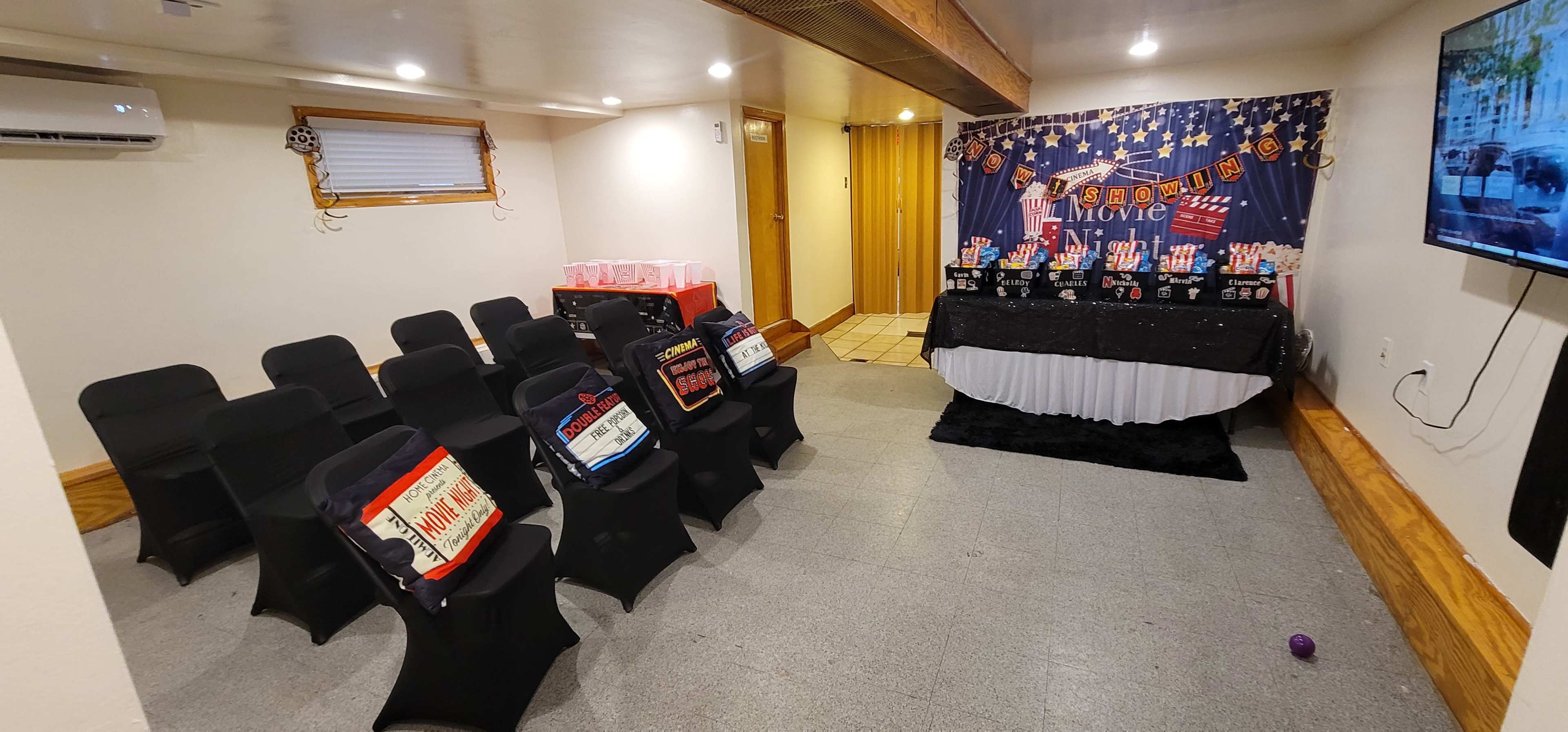 The scene shows a small movie-themed party room with rows of black chairs, a decorated table with a movie backdrop, and a television displaying a movie-themed setup.