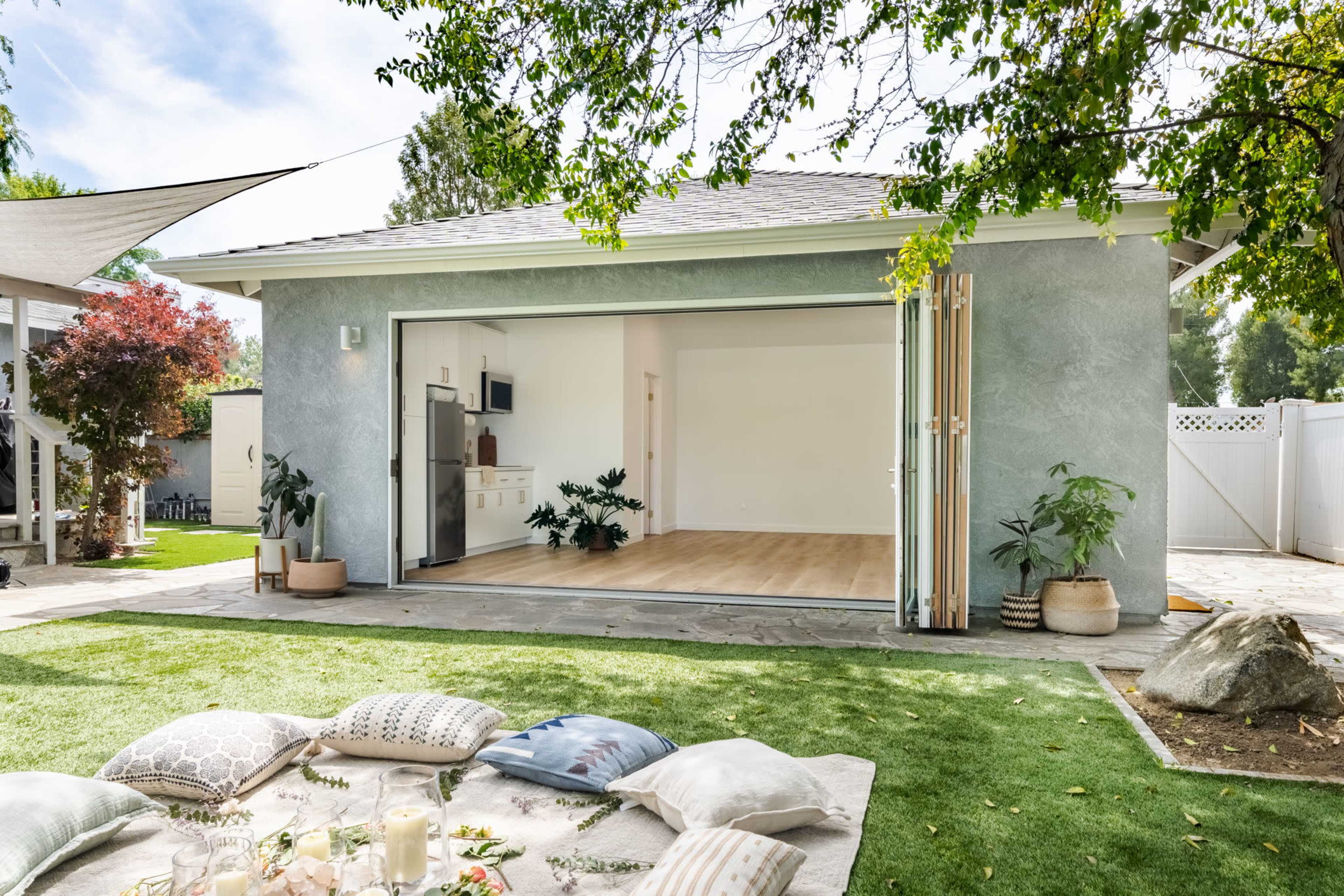 A modern outdoor space features a sliding glass door leading into an open room with wooden flooring, surrounded by greenery and decorative cushions on the grass.