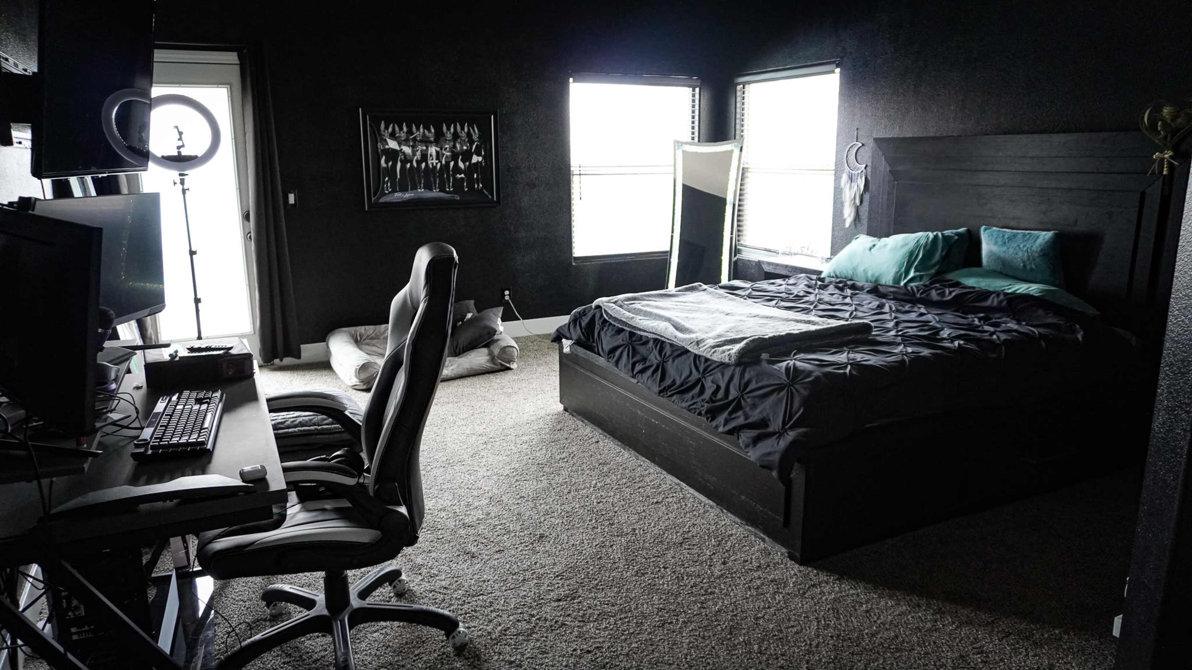 A dark-themed bedroom features a large bed with teal pillows, a gaming desk with a computer, and large windows letting in natural light.