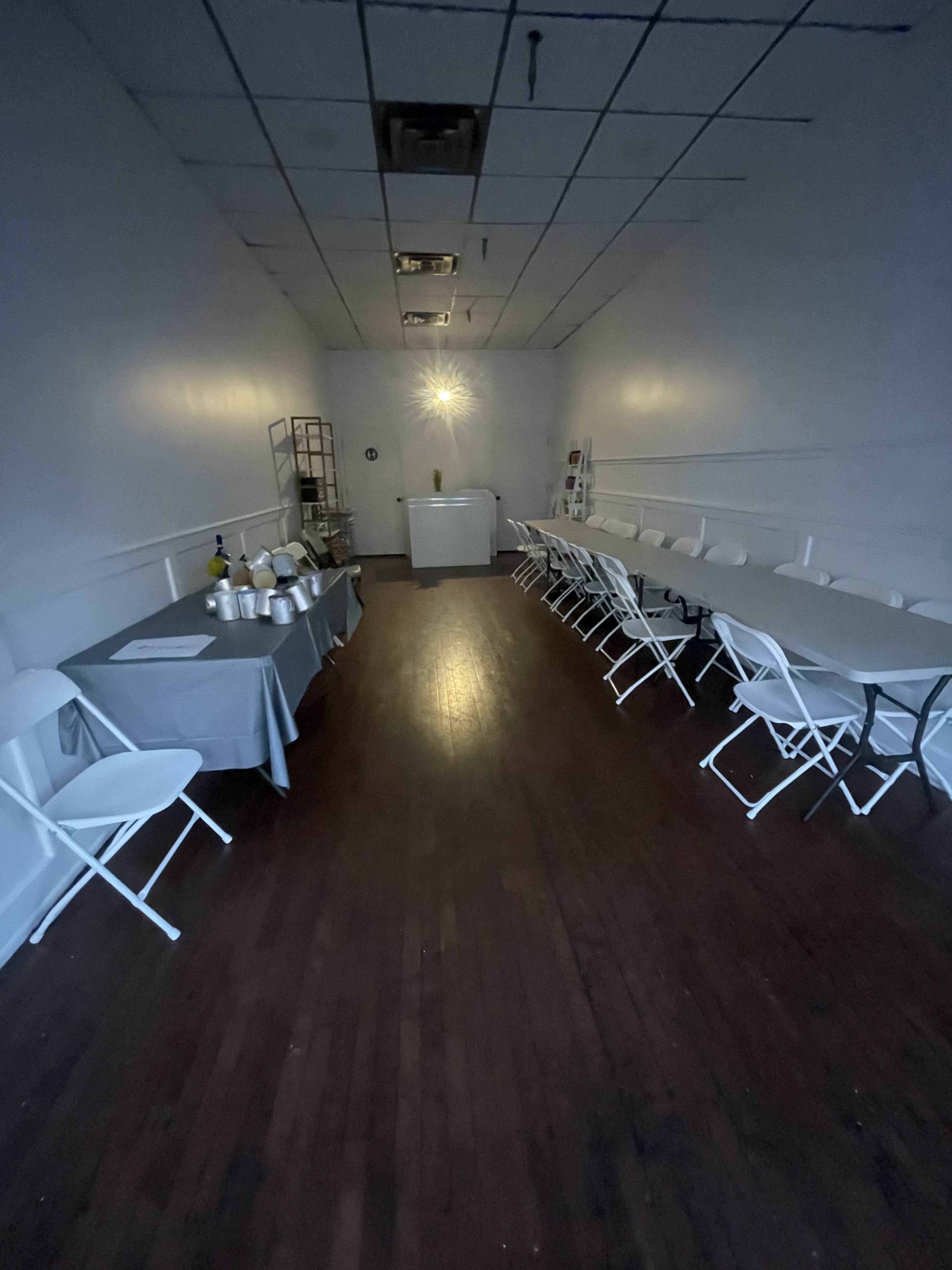 A long, narrow room with white walls features several folding tables and chairs arranged in two rows, with a small counter at the far end and a dim light in the center.