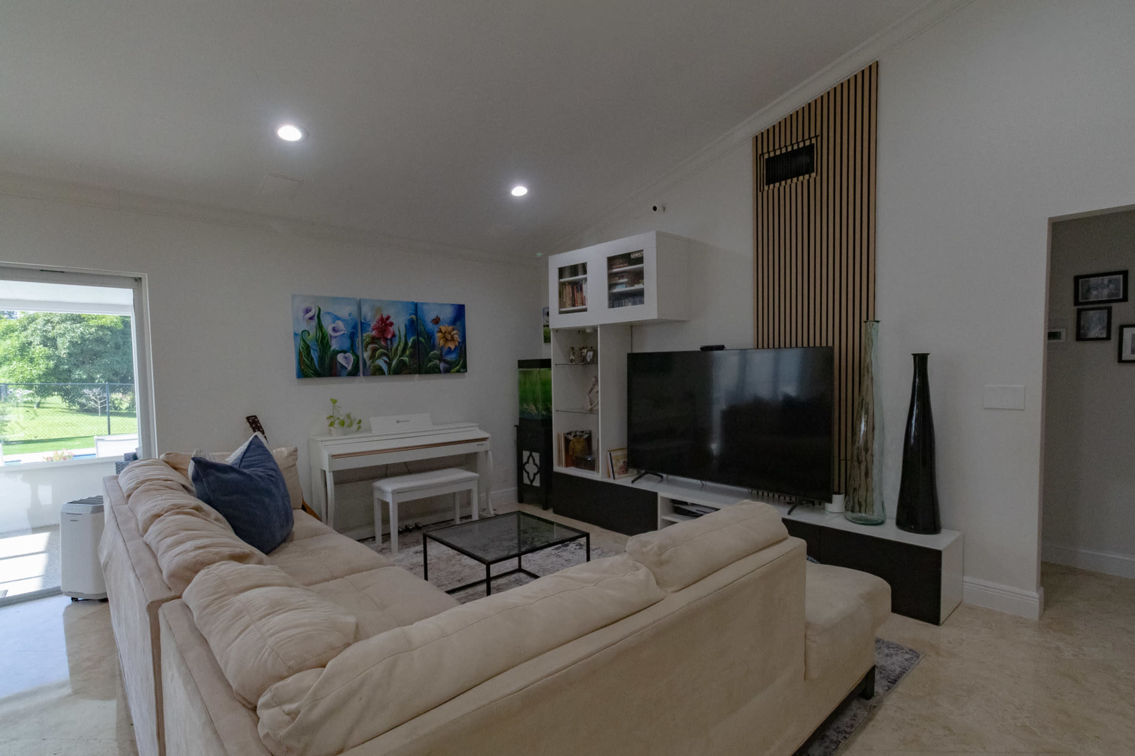 The living room features a beige sectional sofa, a large flat-screen TV, a white piano, and decorative furniture against a neutral backdrop with large windows.