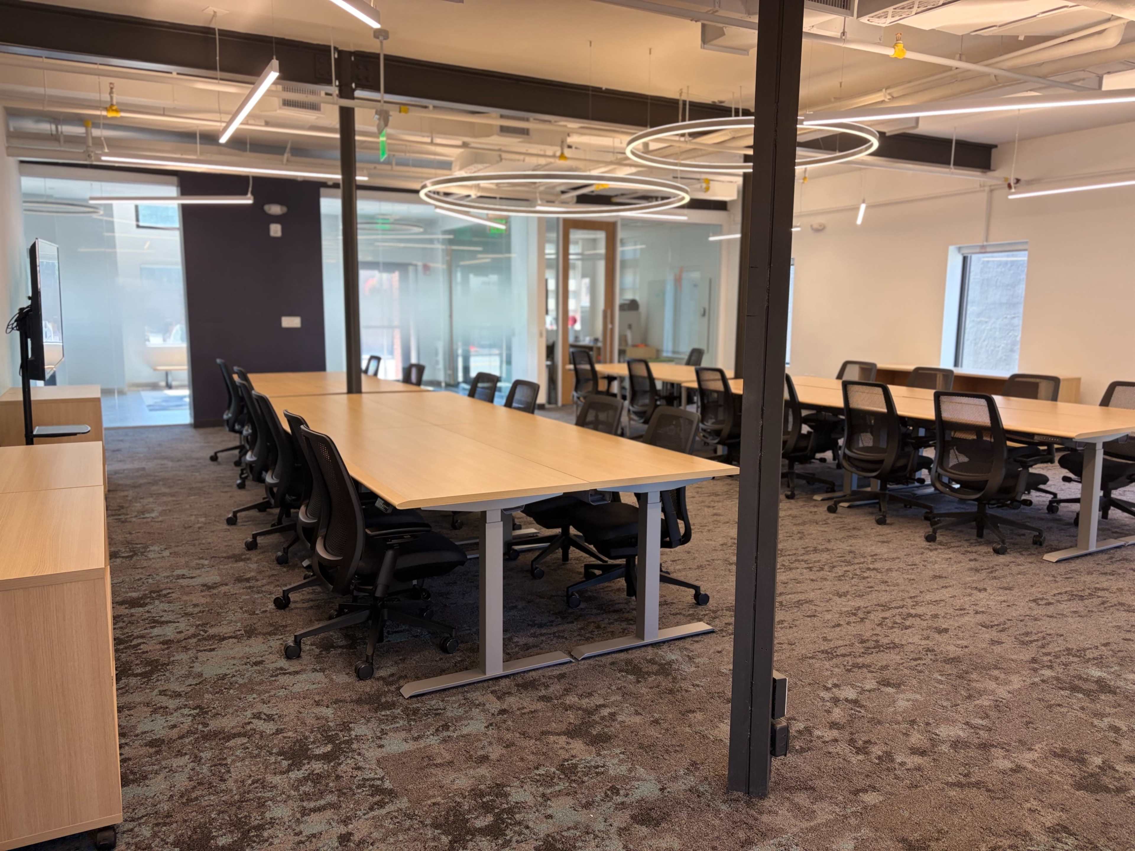The image shows a modern office space with large tables and ergonomic chairs arranged in a well-lit environment.