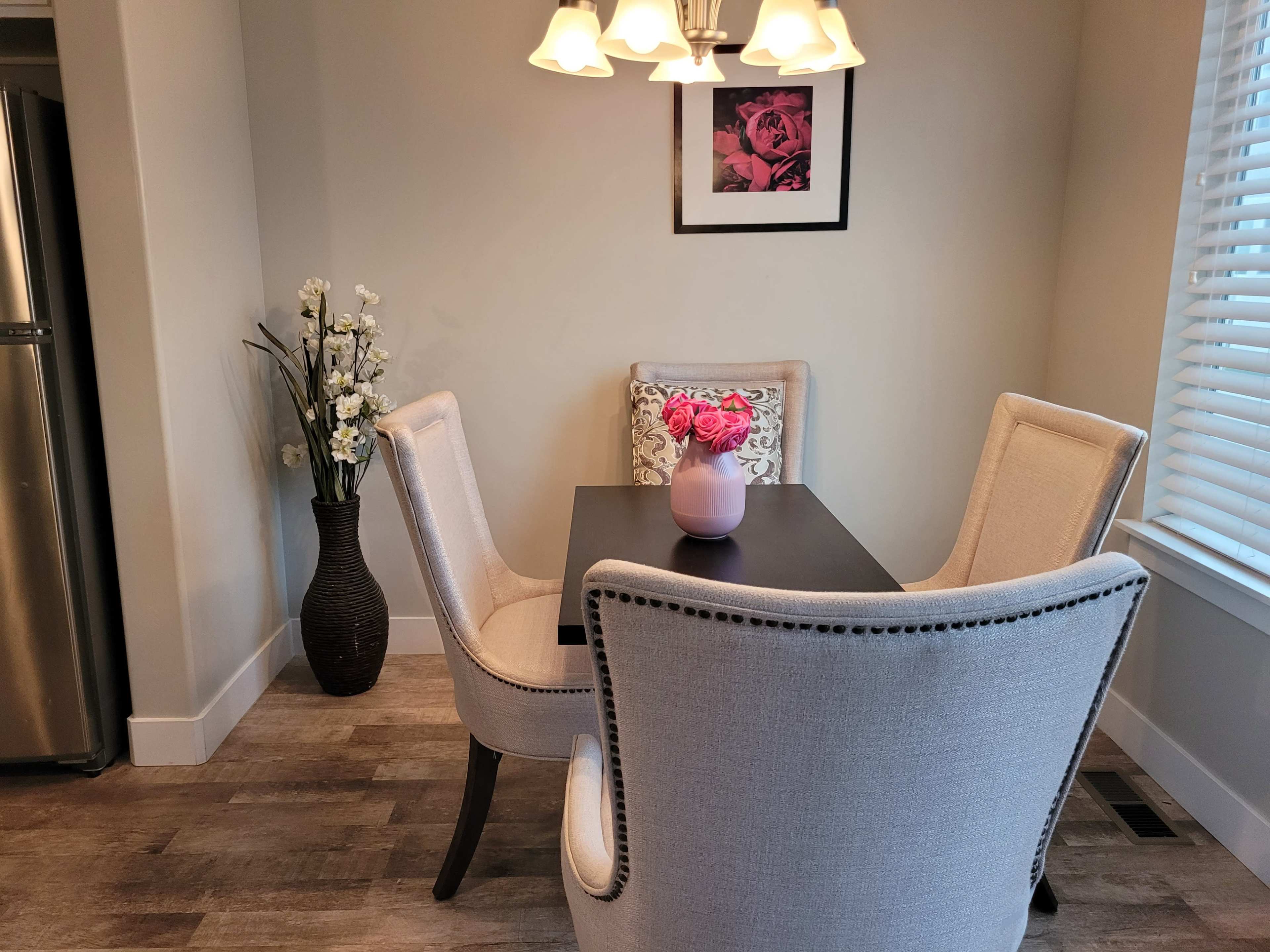 A small dining area features a black table surrounded by two upholstered chairs, a vase of pink flowers on the table, and a wall with a framed picture and a decorative plant.