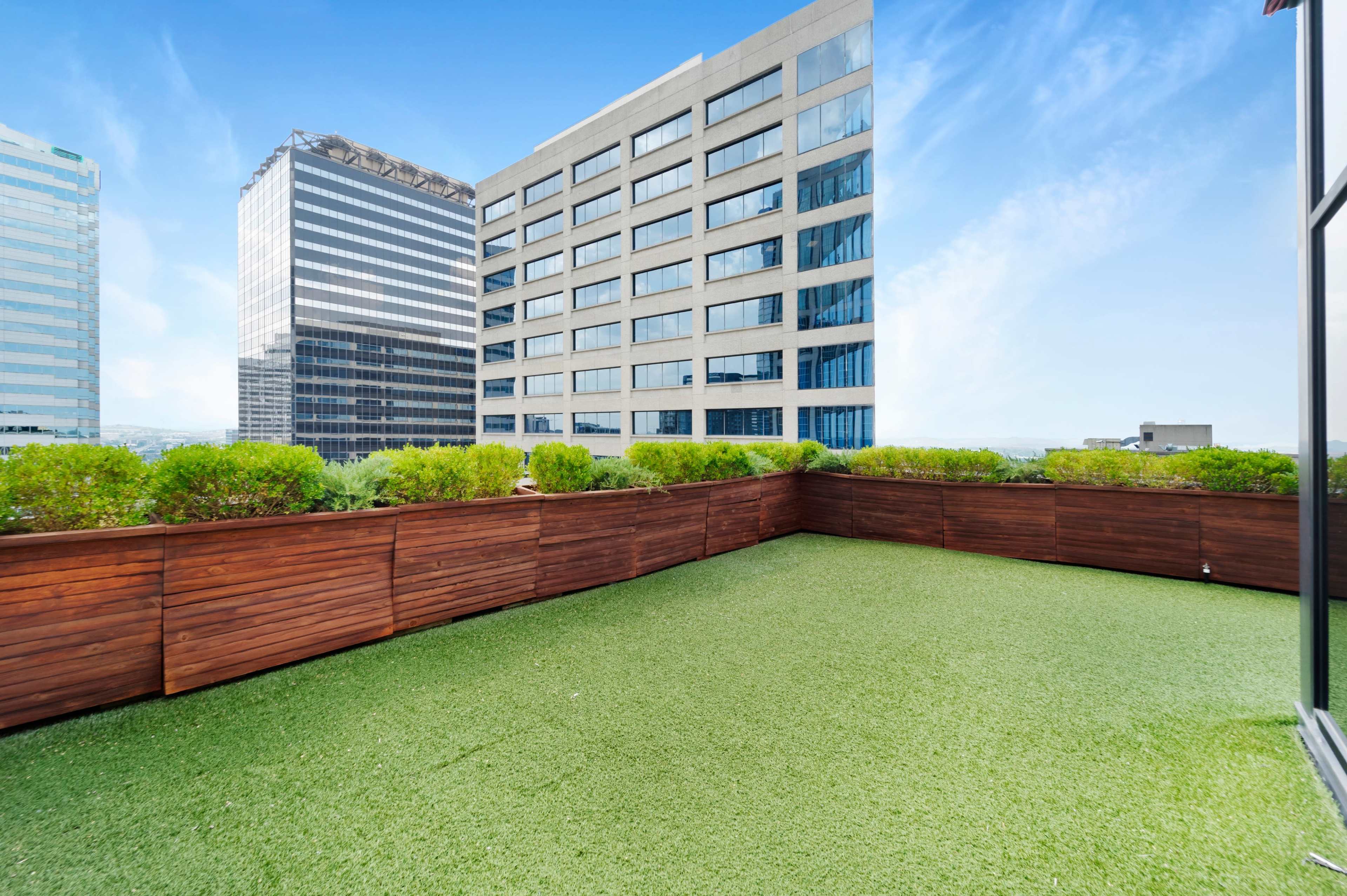 A rooftop terrace with artificial grass and wooden fencing, overlooking adjacent high-rise buildings.