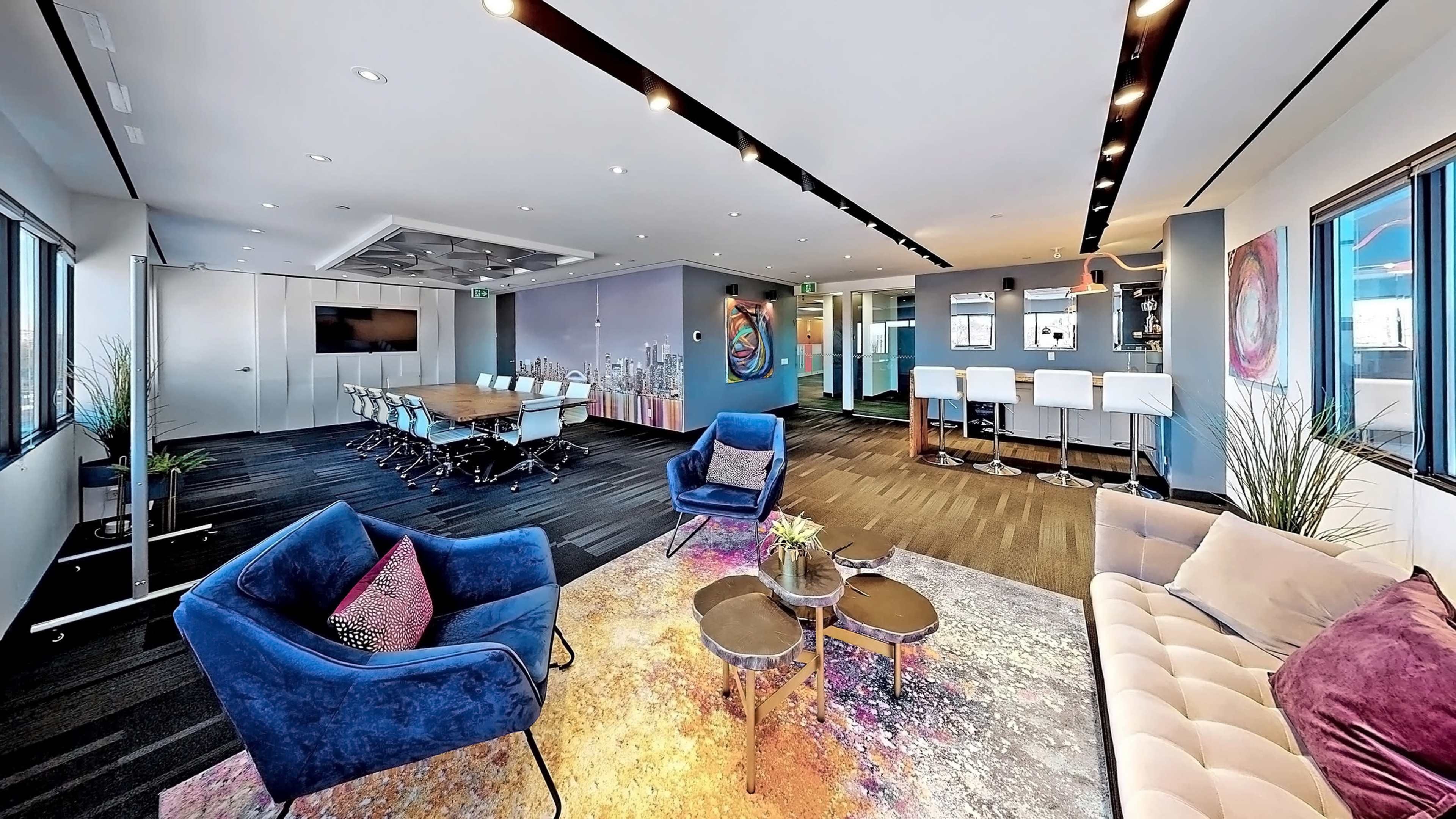 The image shows a modern office space featuring a meeting area with a large table, lounge seating with blue chairs, and a bar area with stools.
