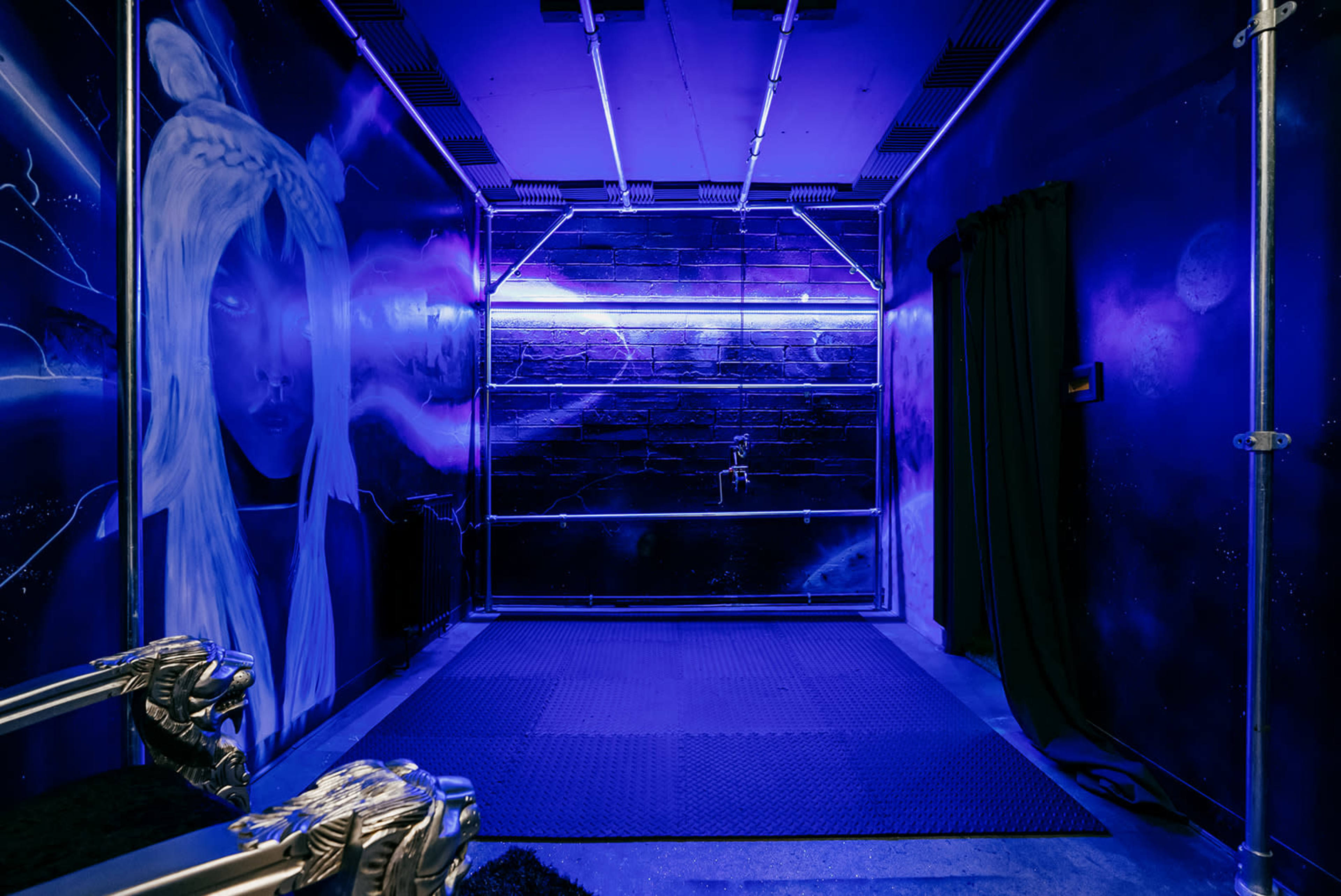 A dark room with purple lighting, featuring a mural of a woman on one wall and a curtain on another.