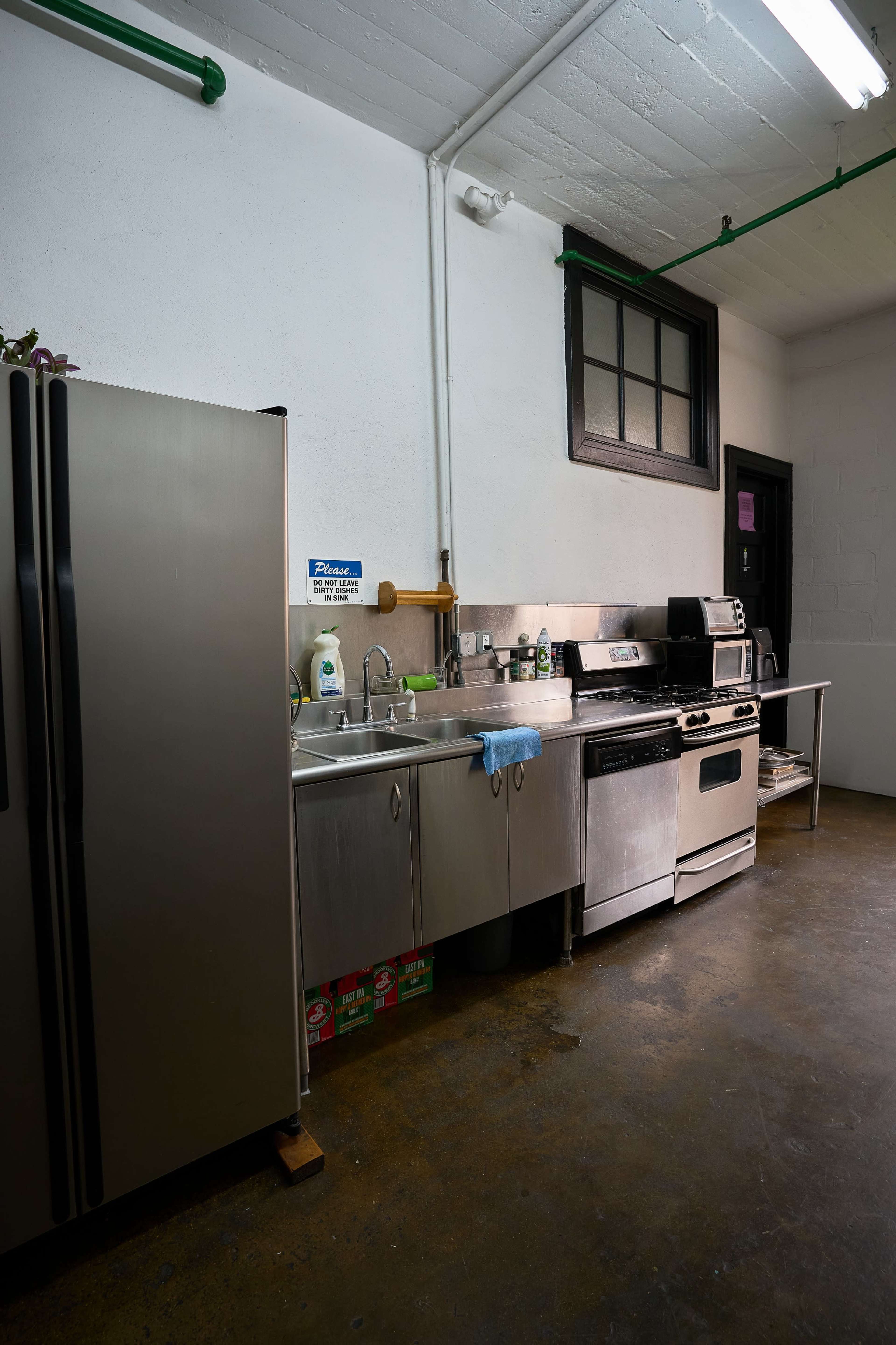 A kitchen with stainless steel appliances, including a refrigerator, sink, stove, and countertop, situated against a concrete wall.
